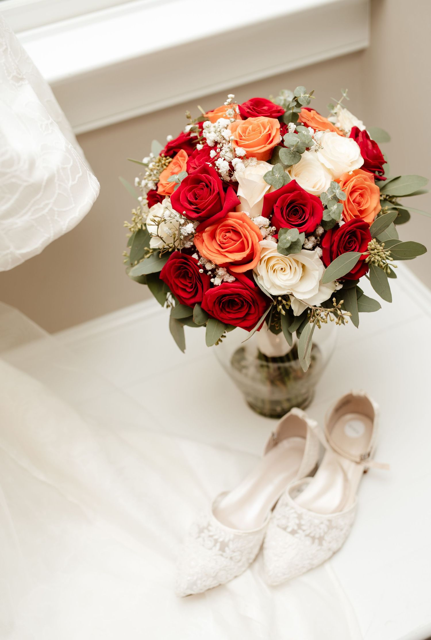 Wedding detail shots of red and peach roses in a vase next to bridal shoes on a white surface.