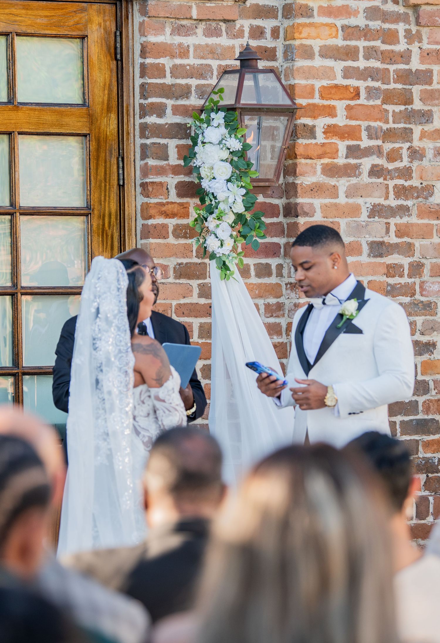Wedding party members stand in white and black formal attire against brick backdrop.