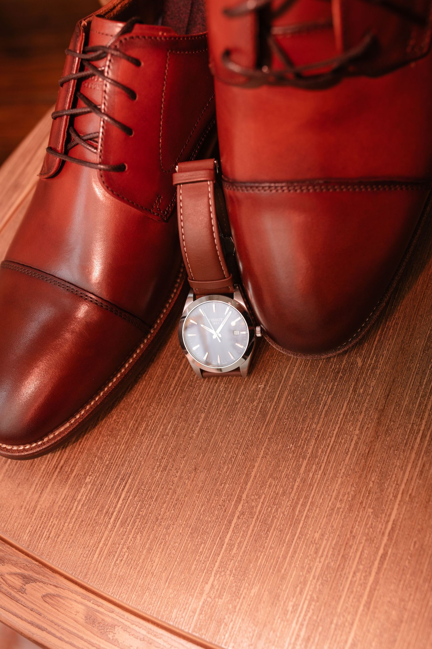 Brown leather dress shoes and silver watch arranged on wooden surface for groom's wedding accessories.