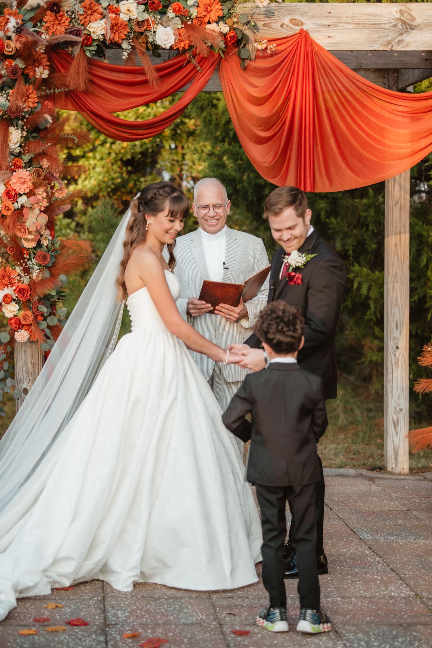 Wedding ceremony participants stand beneath a rustic wooden arch with coral draping and flowers.