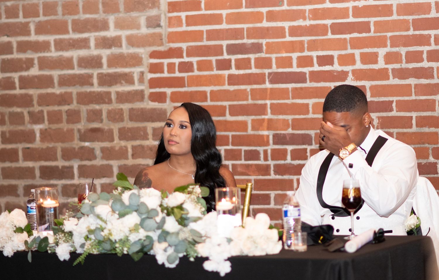A formal reception table setting with elegant floral centerpieces against a brick wall backdrop.
