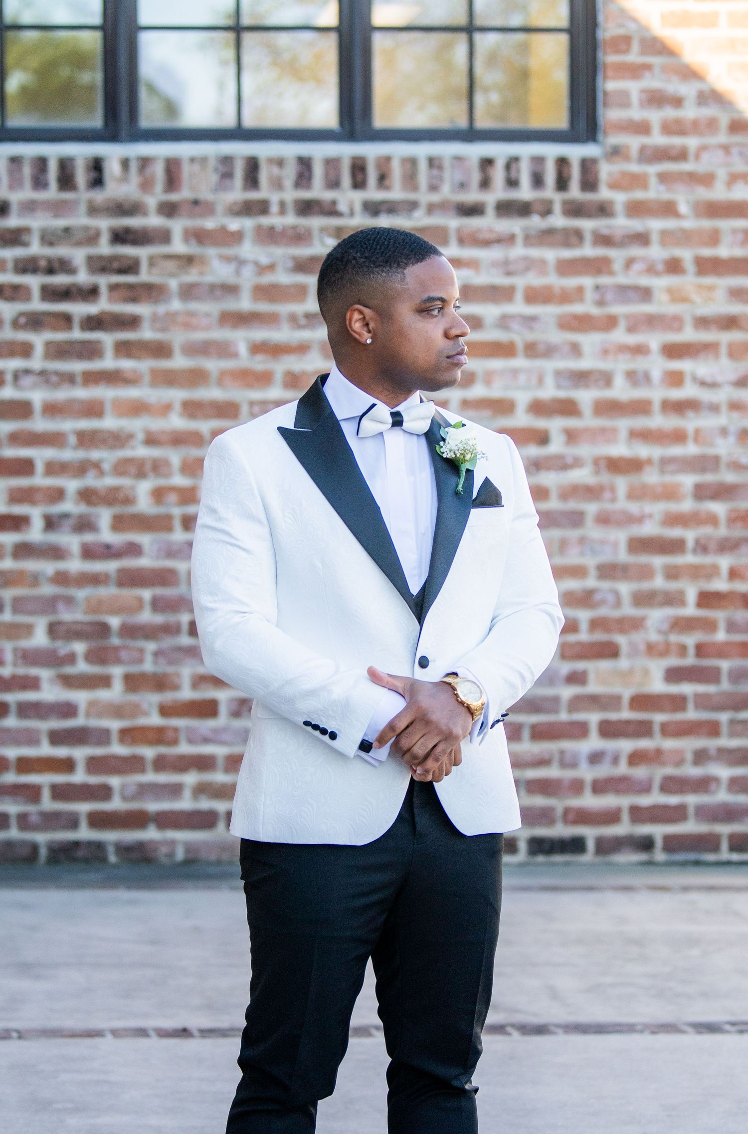 Groomsman in white tuxedo jacket and black pants poses against brick wall.