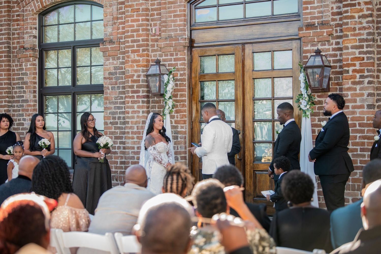 Wedding ceremony audience watches celebration in front of rustic brick building facade.