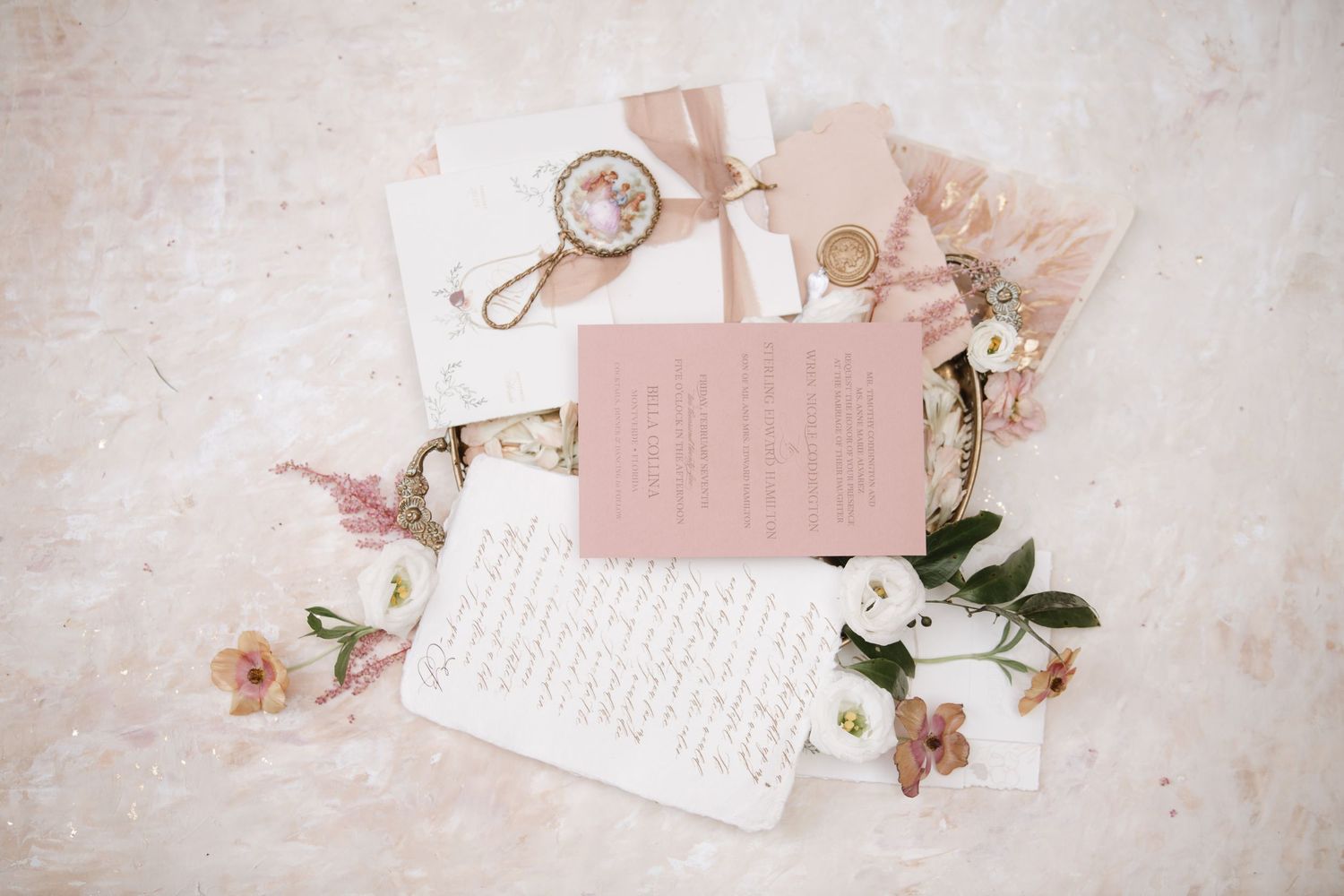 Romantic flat lay of pink wedding stationery with floral accents on white textured background.
