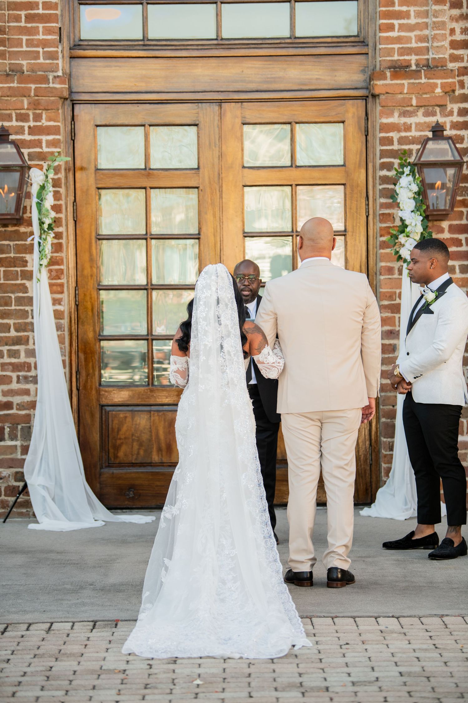 Wedding party members interact outside historic brick venue with wooden doors and windows.