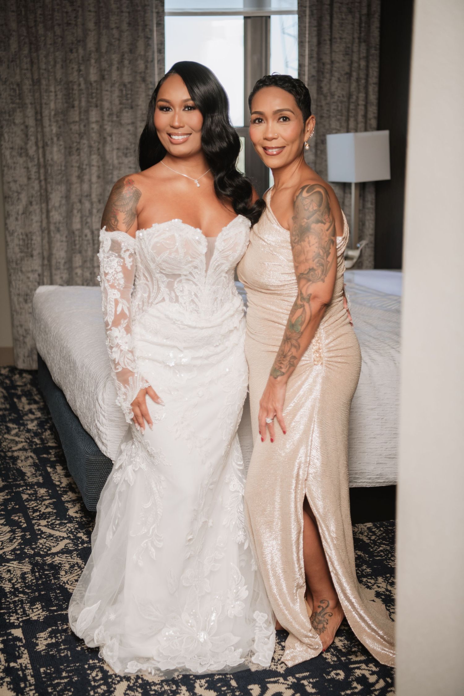 Two people in contrasting white and champagne wedding dresses pose together on a patterned carpet.