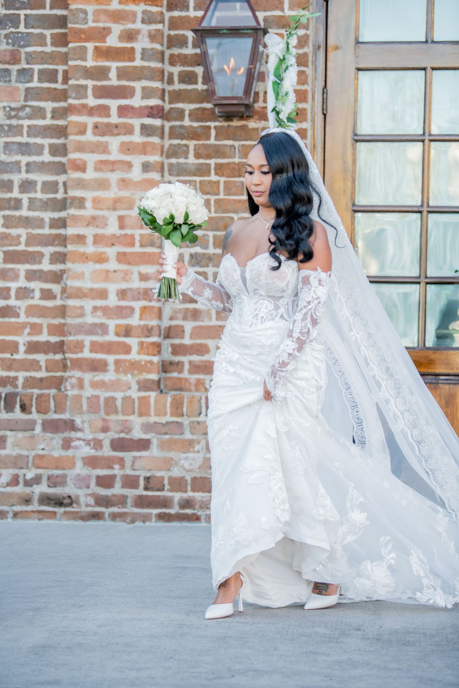 Bride in elegant lace wedding gown poses gracefully against rustic brick wall background.