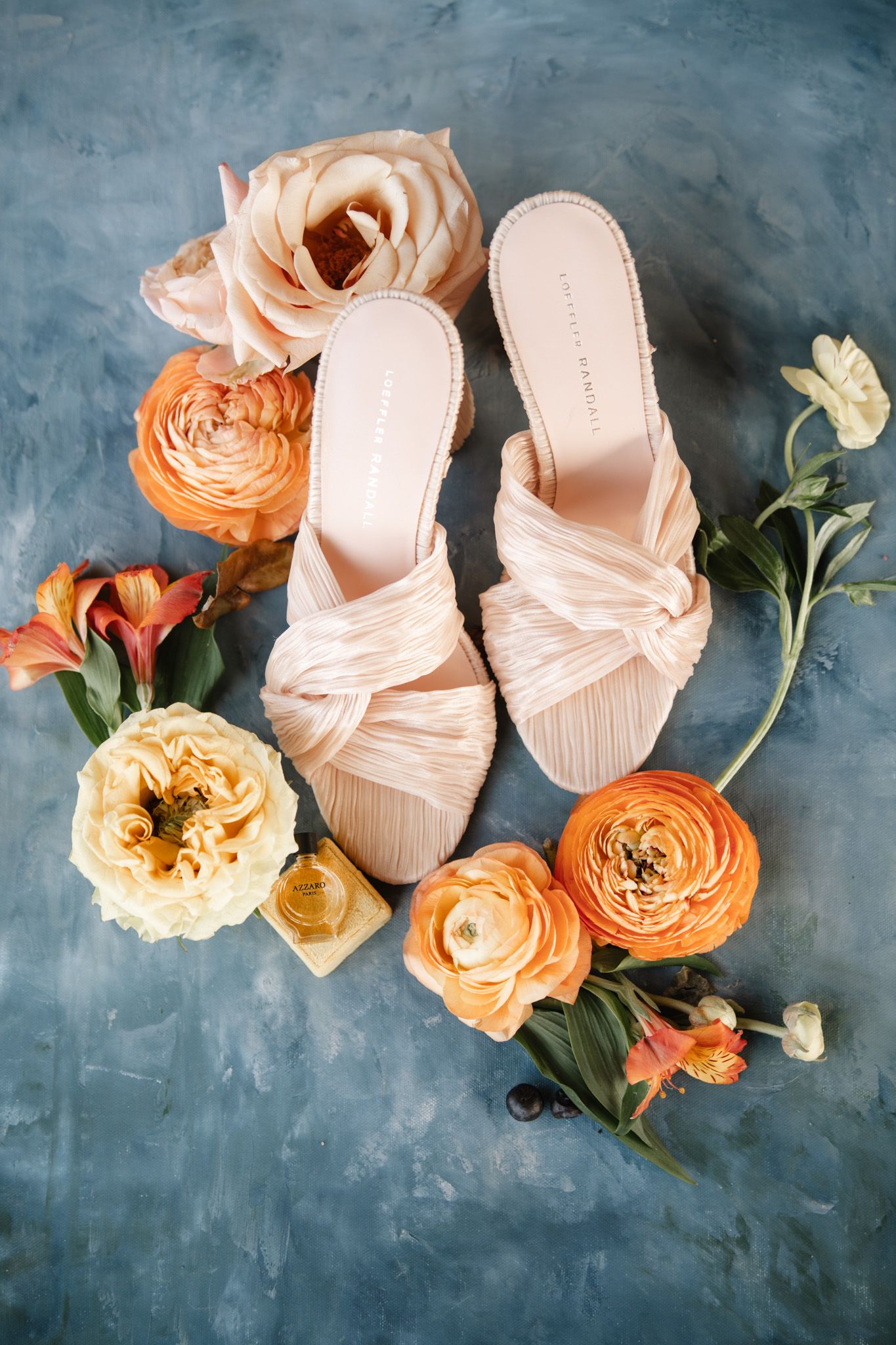 Blush pink bridal shoes with peach ranunculus flowers on blue textured background.