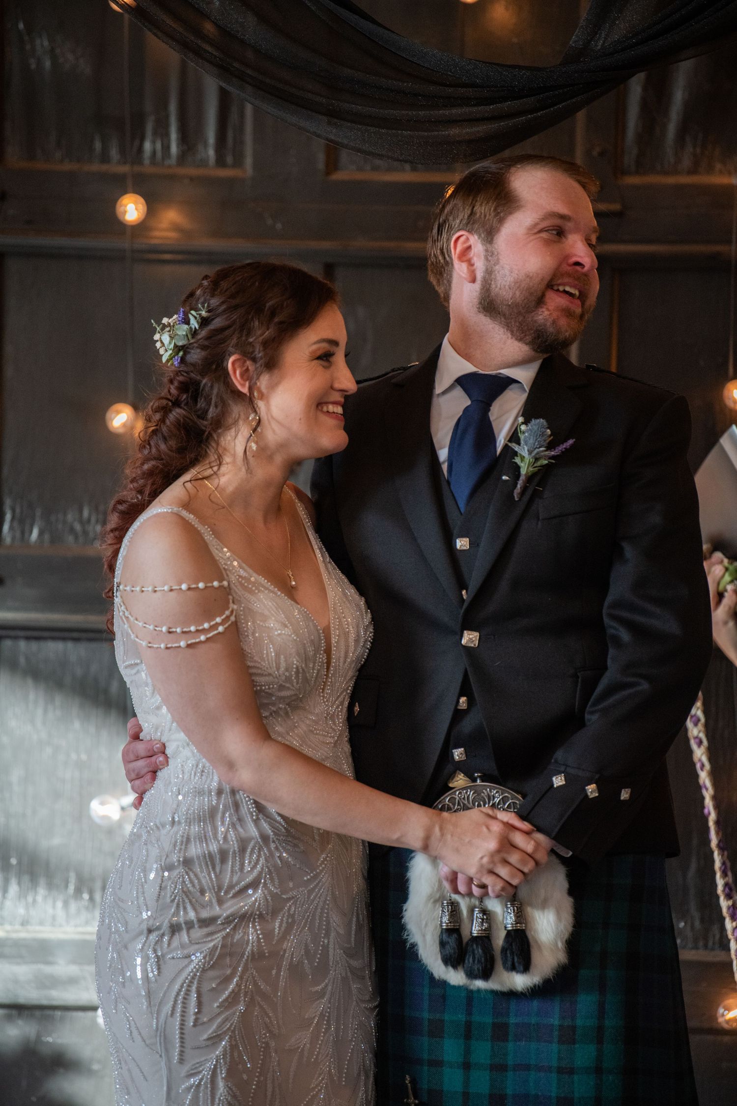 Newlyweds sharing a joyful moment in venue decorated with string lights and dark draping.