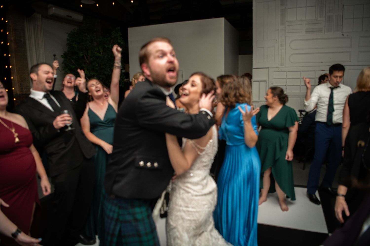Wedding guests dancing energetically at a reception with colorful formal attire and kilts.