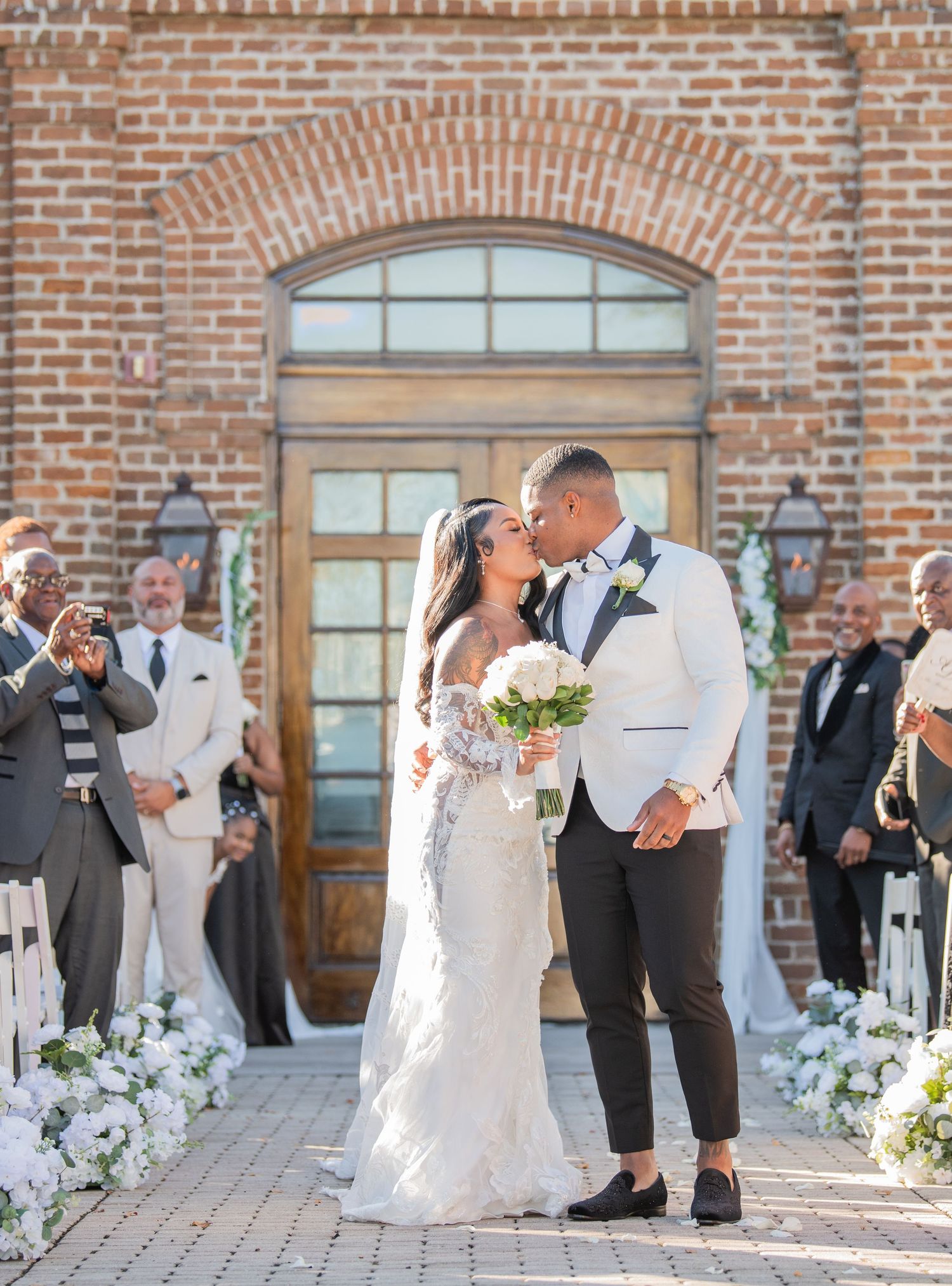 Wedding couple poses for photos outside brick church with wedding party looking on.