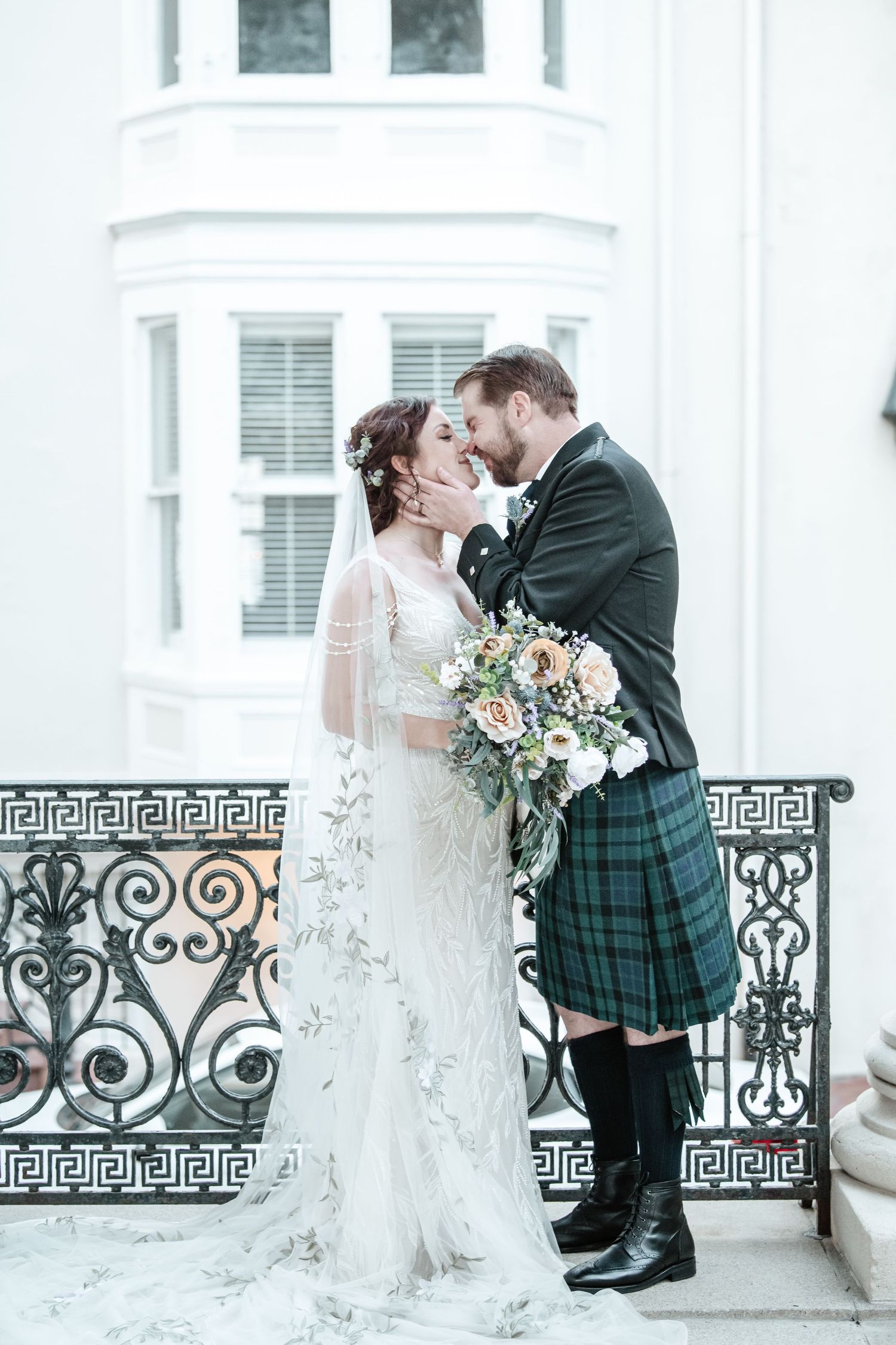 Wedding couple embracing by decorative iron railing against white building backdrop.