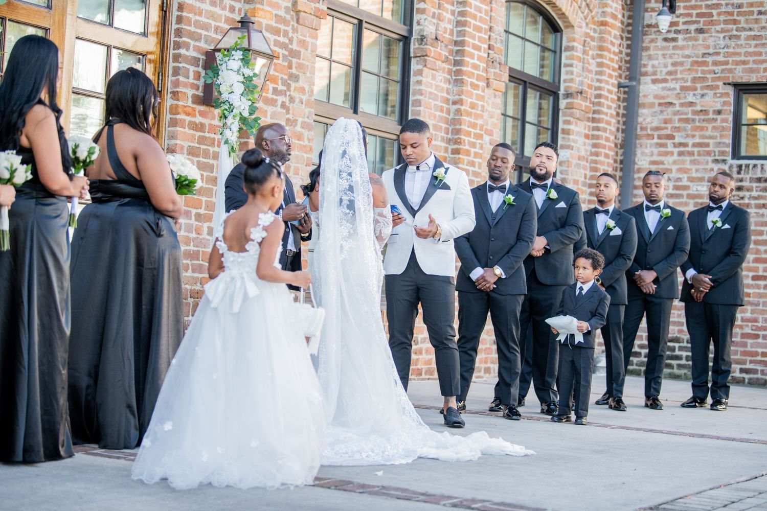 Wedding ceremony participants line up along brick wall with decorative white drapery.