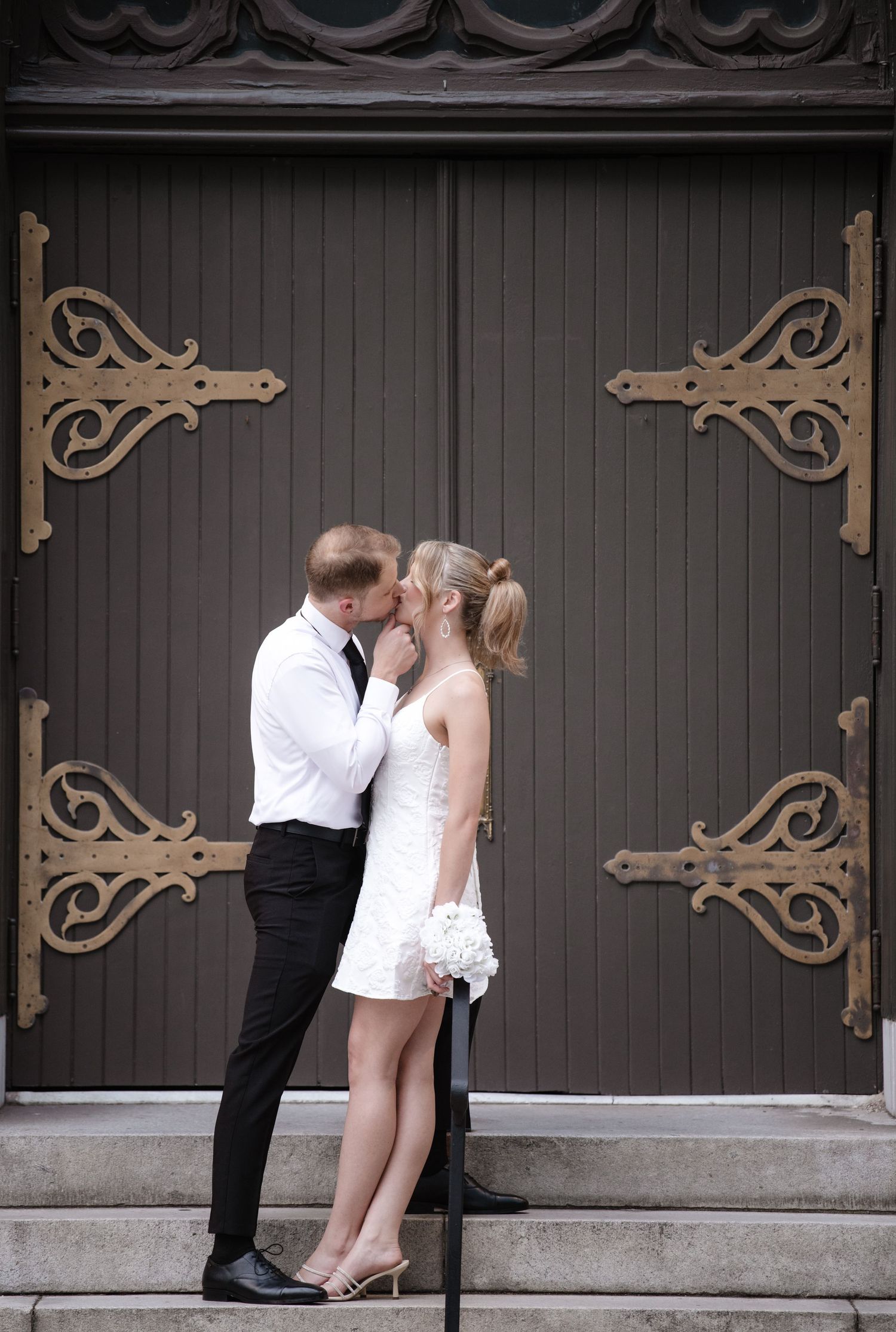 A couple shares an intimate moment in front of a decorative dark wooden church door.