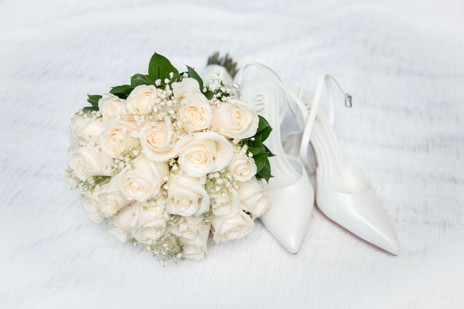 White wedding shoes arranged with cream roses and greenery on textured background.