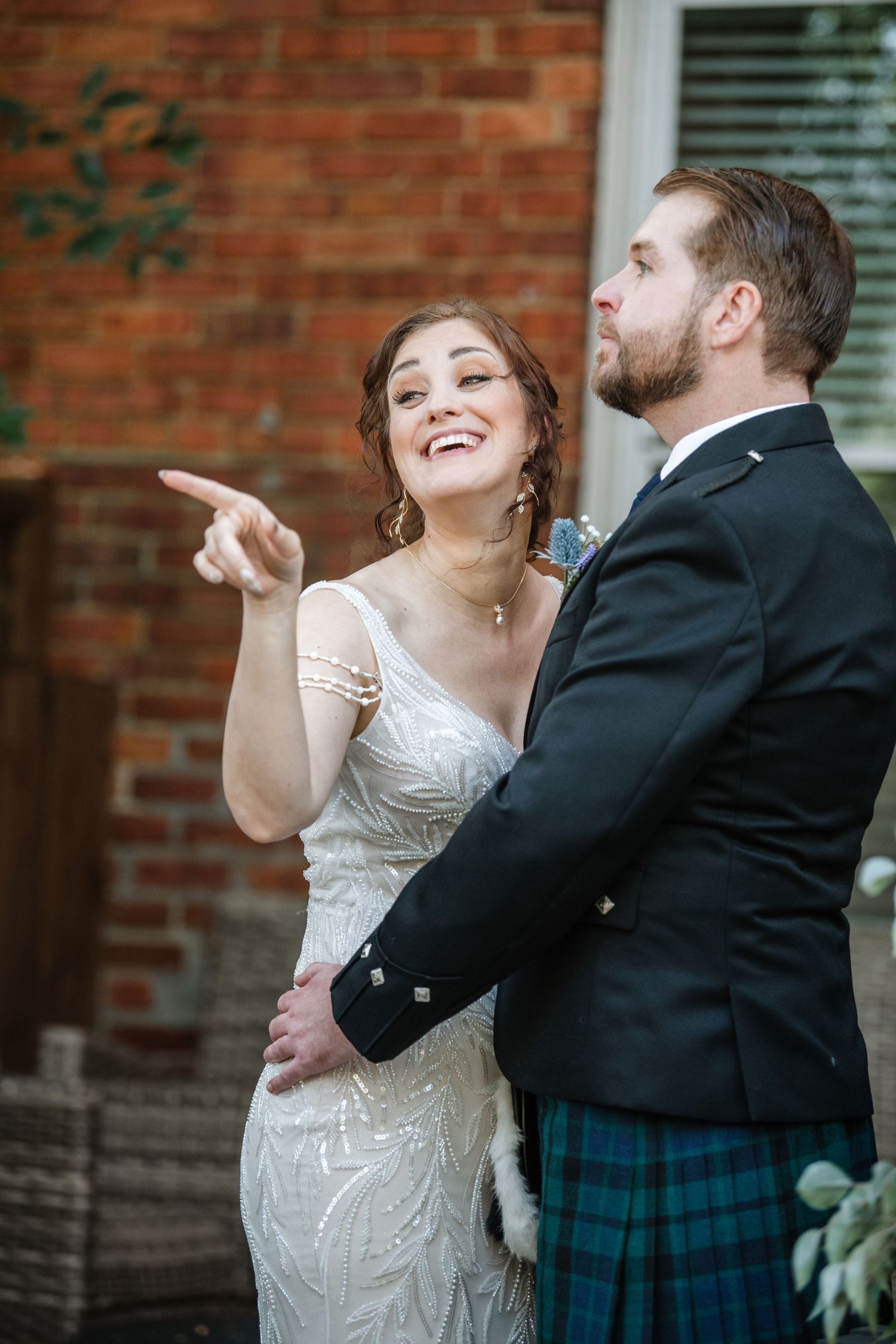 Playful moment between couple as bride points and laughs while groom wears traditional Scottish formal wear.