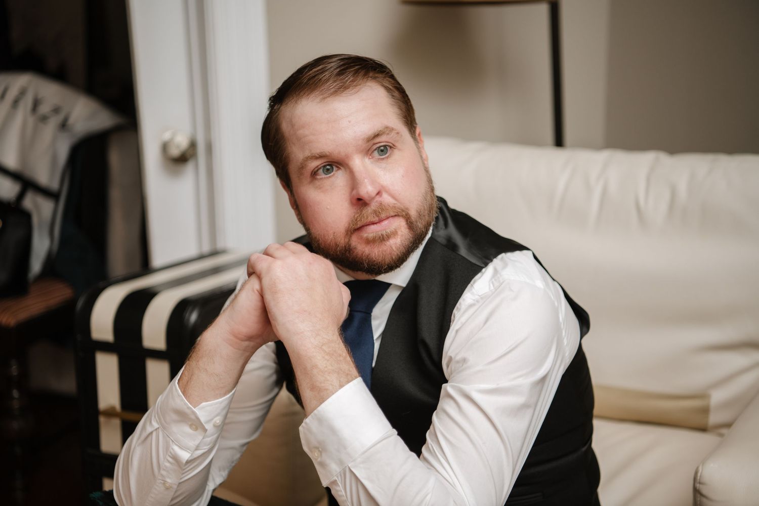 A person in formal attire sits thoughtfully in a living room setting.