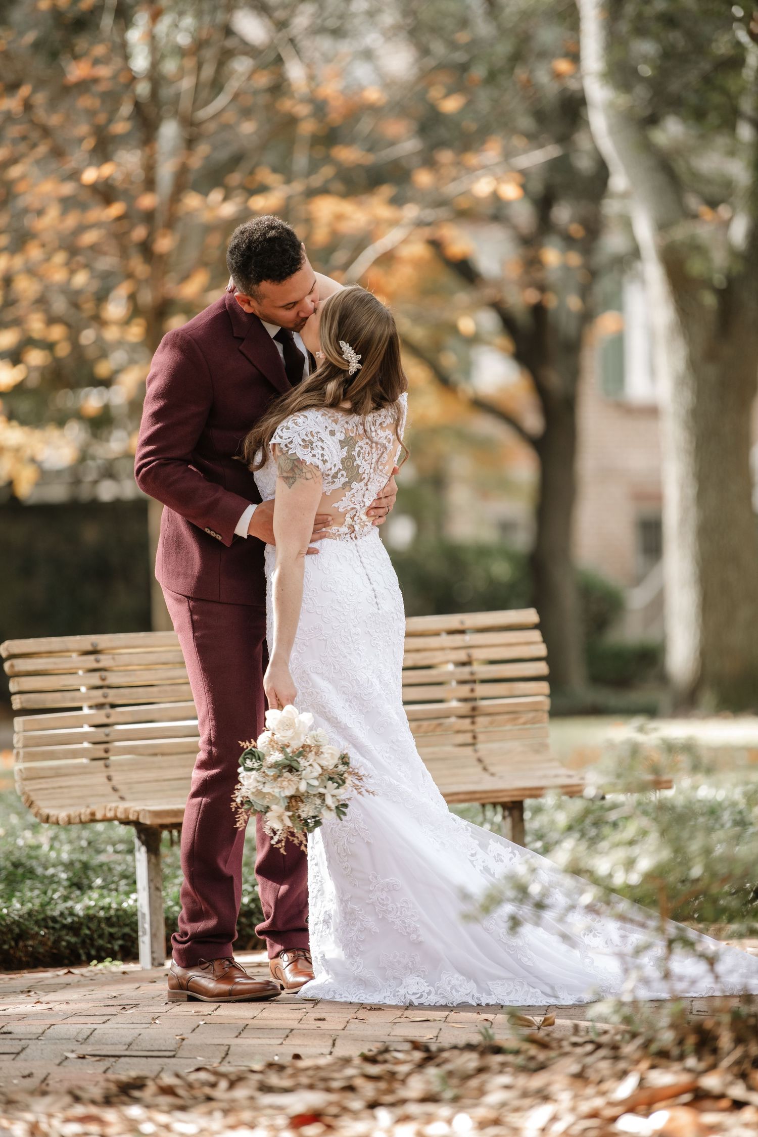 Newlyweds share intimate moments on wooden bench during autumn wedding photoshoot.