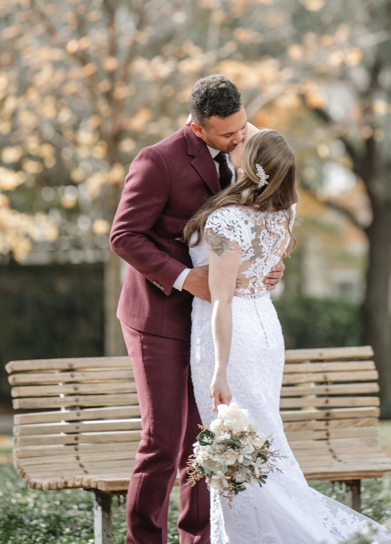 A couple in burgundy suit and white lace dress share an intimate moment on a park bench.