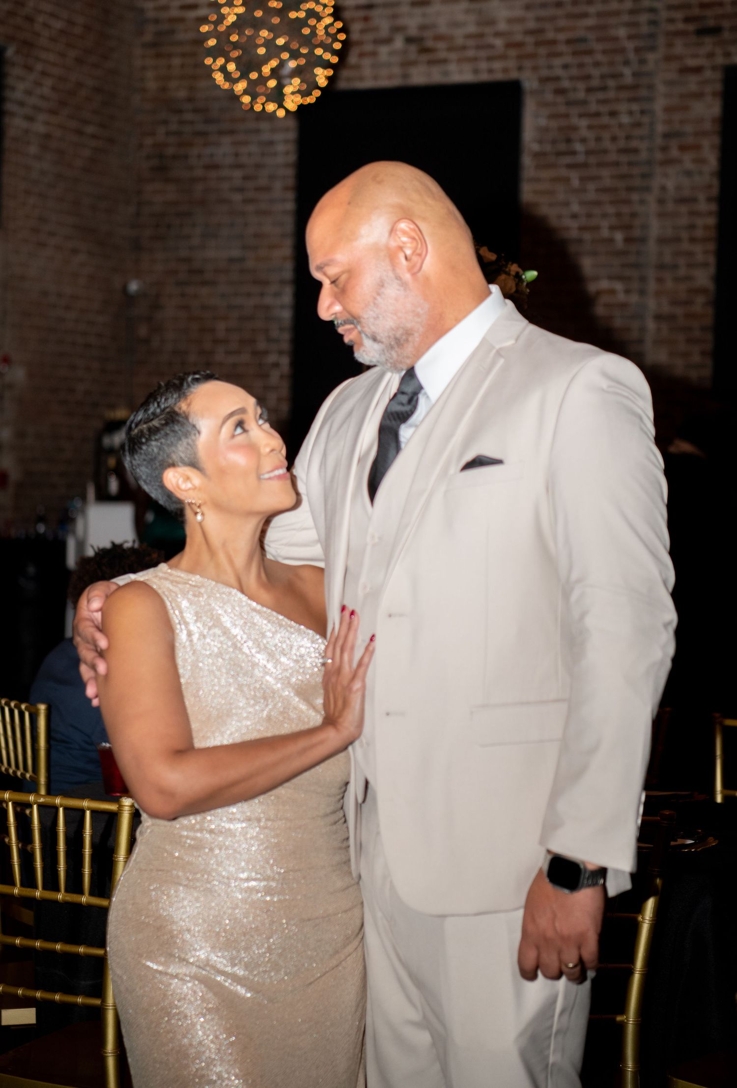 Couple in sparkly gold dress and white suit dance together at wedding reception.