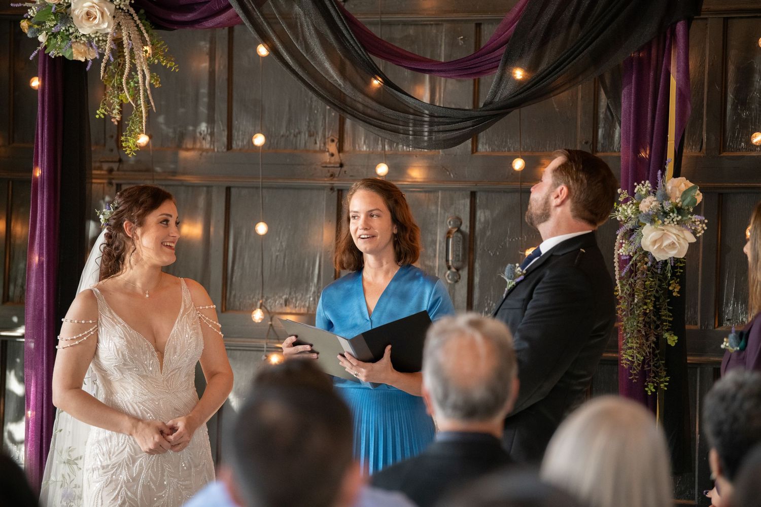 Wedding ceremony in a rustic venue with string lights and purple draping decor.