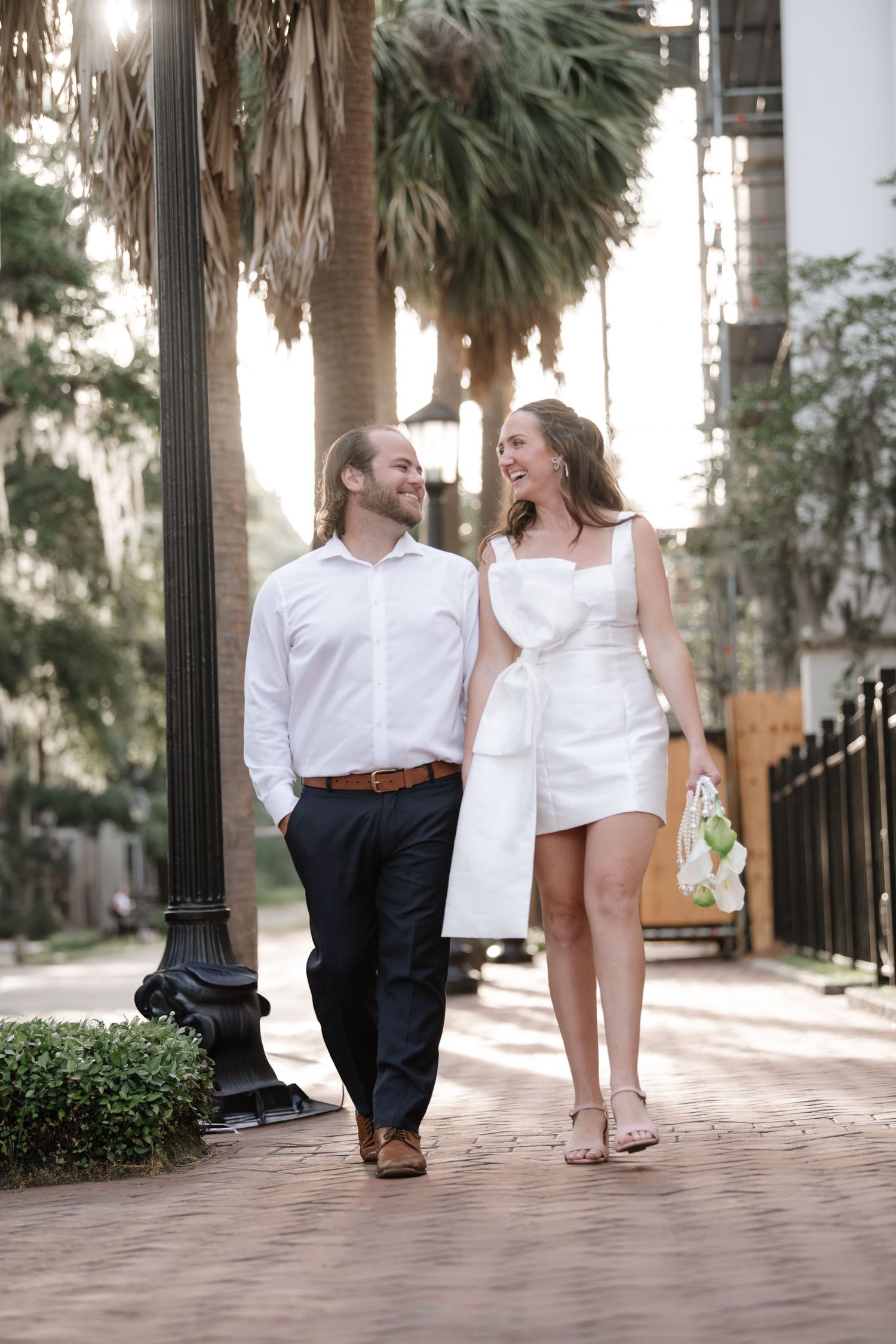 A couple in white and black outfits walks together on a brick pathway lined with palm trees.