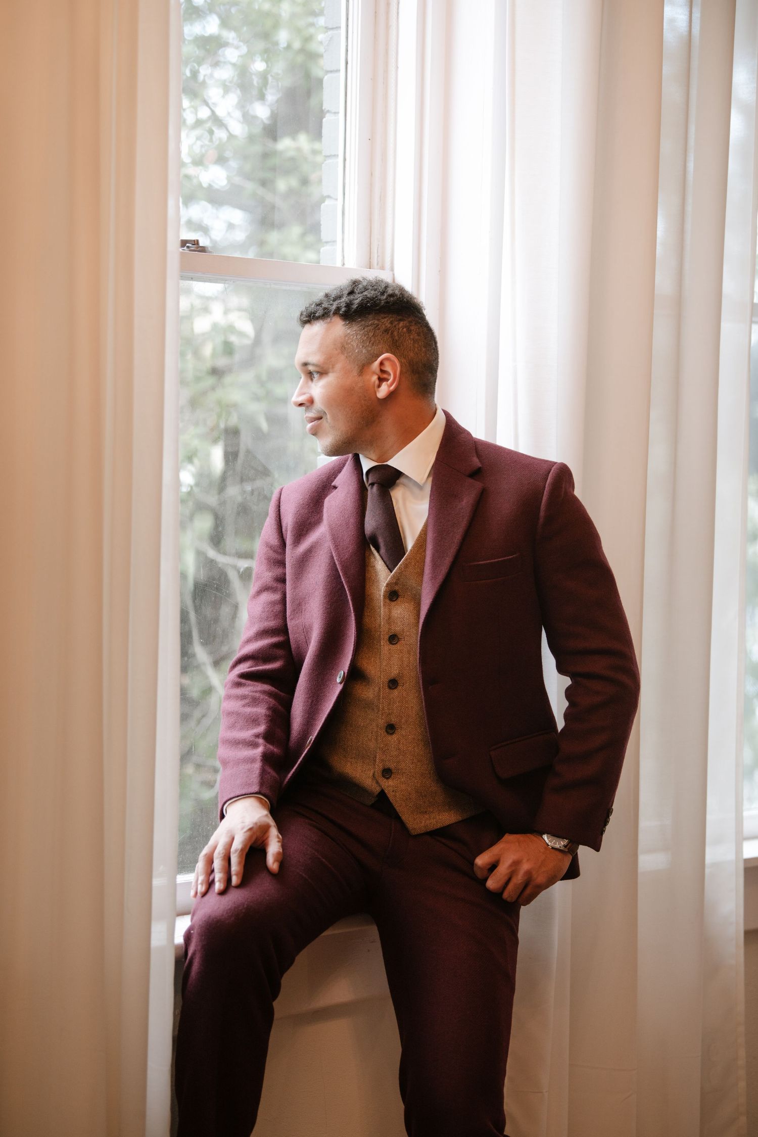 Elegant burgundy suit with brown vest styled against bright window in formal setting.