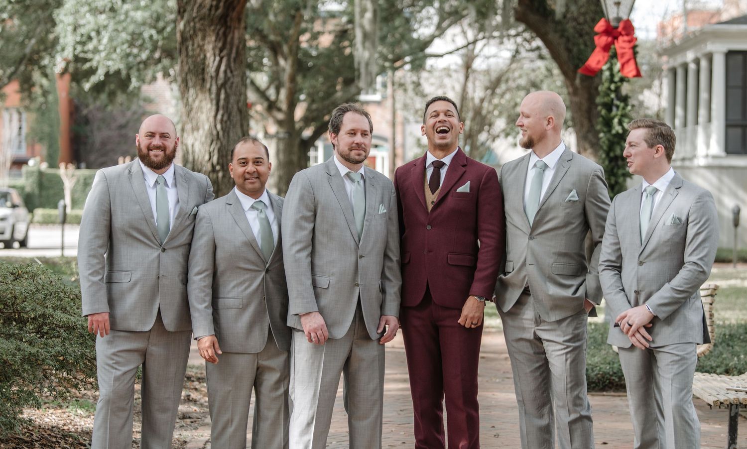 Groomsmen in grey suits stand with one member wearing a burgundy suit.
