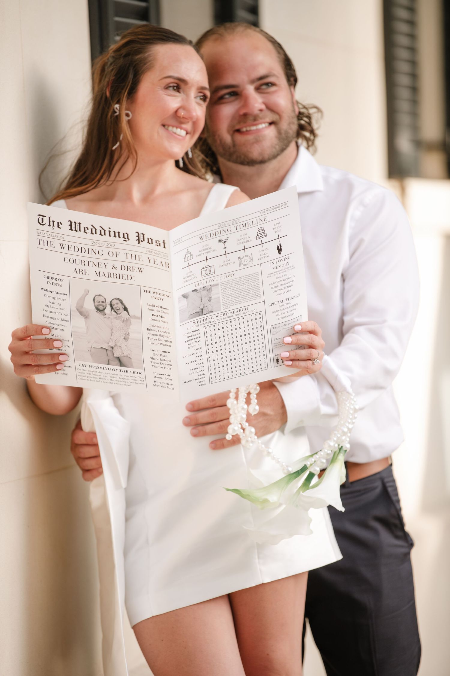 A couple in white clothing poses together while holding up a vintage-style newspaper wedding announcement.