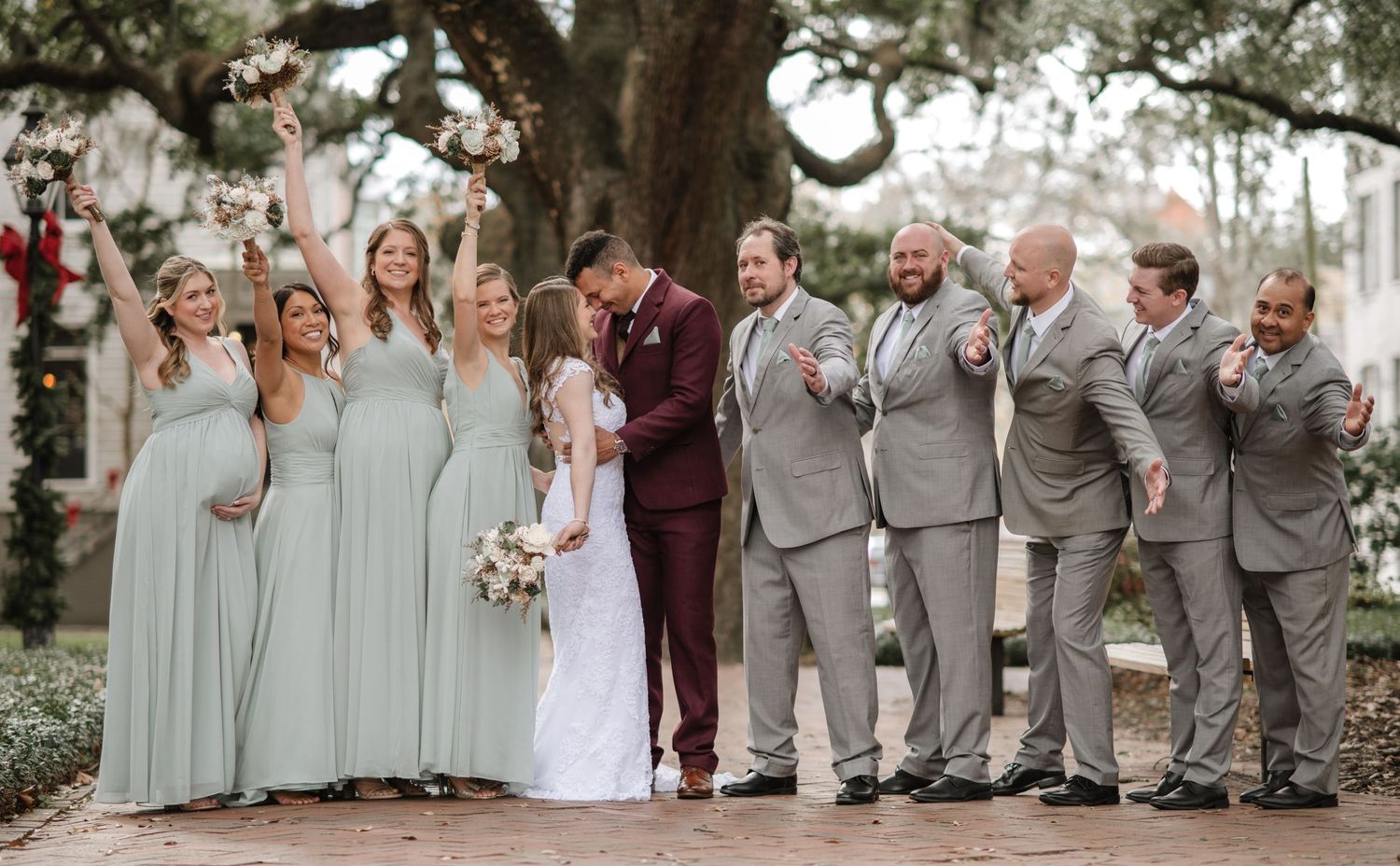 Wedding party in sage green dresses and burgundy suits poses under oak trees.