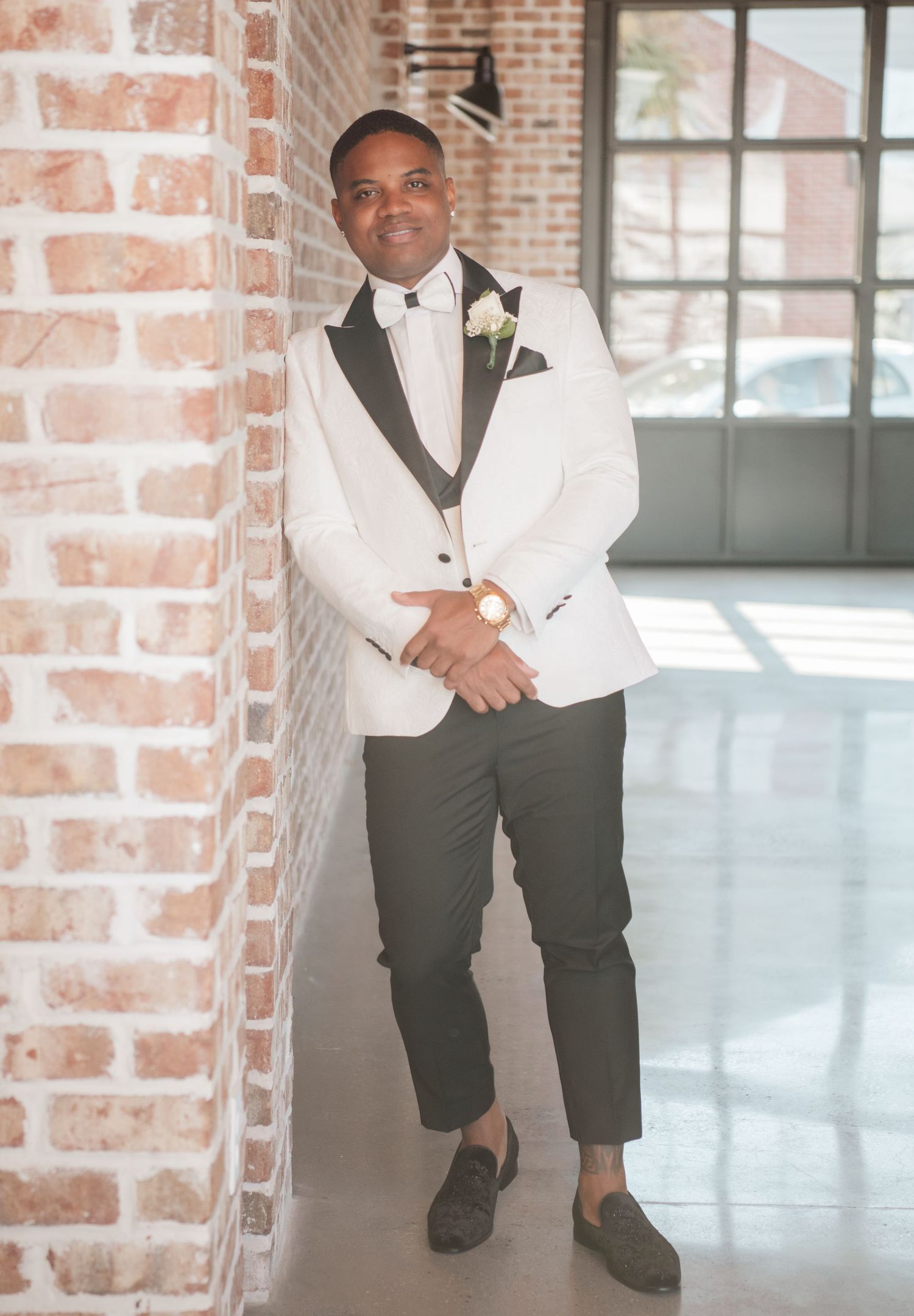 Groomsman in white tuxedo poses against exposed brick wall in natural light.