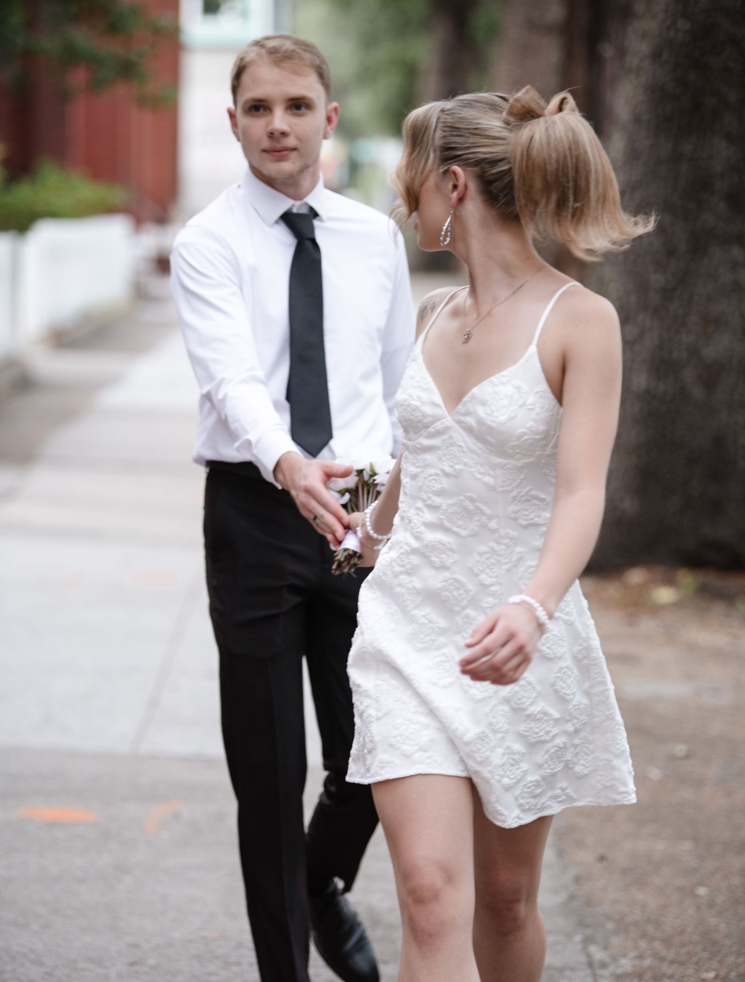 Couple in wedding attire dance playfully on tree-lined street.