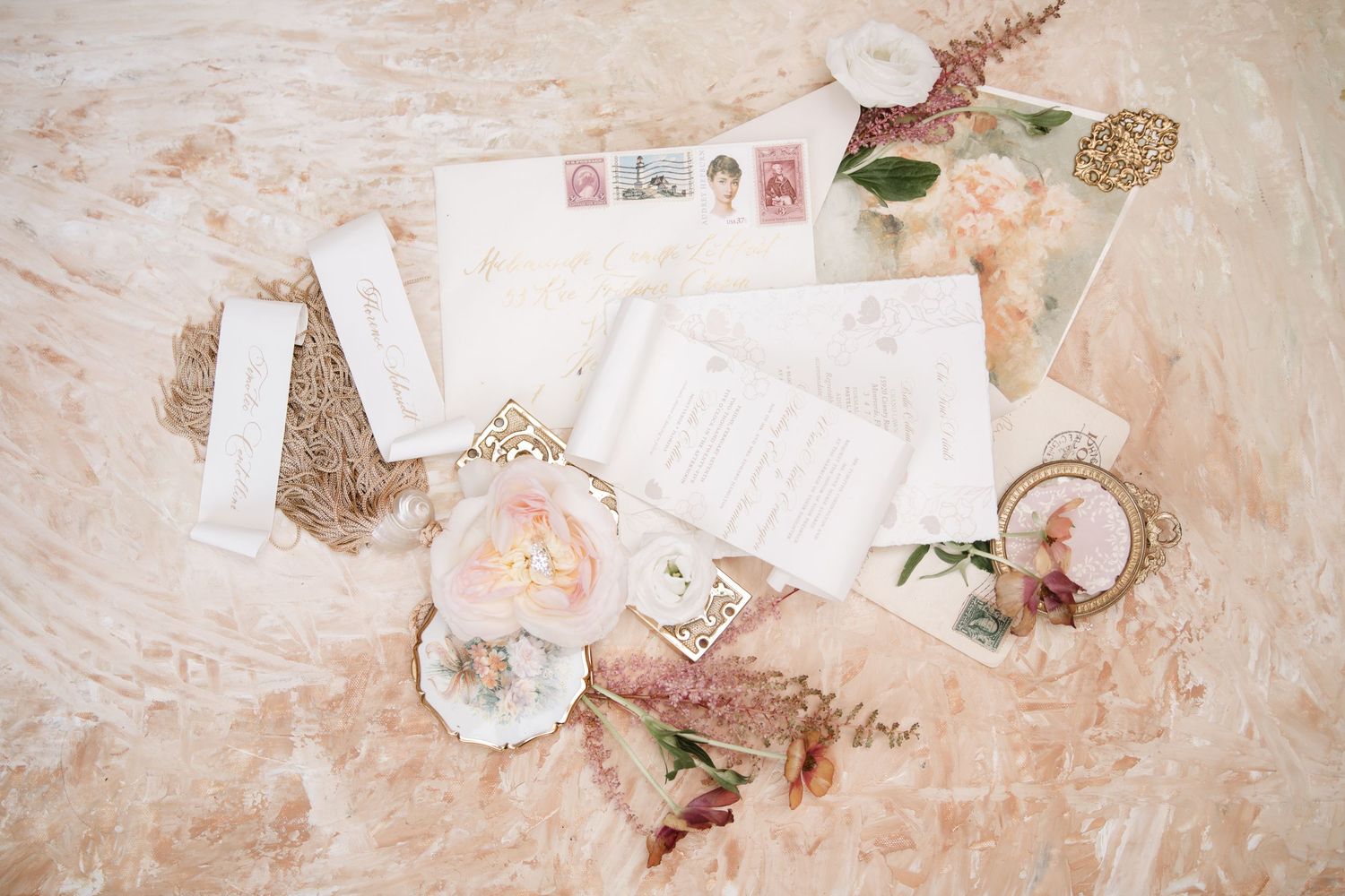 Wedding details flat lay featuring vintage stamps, flowers, and delicate paper elements in pink tones.
