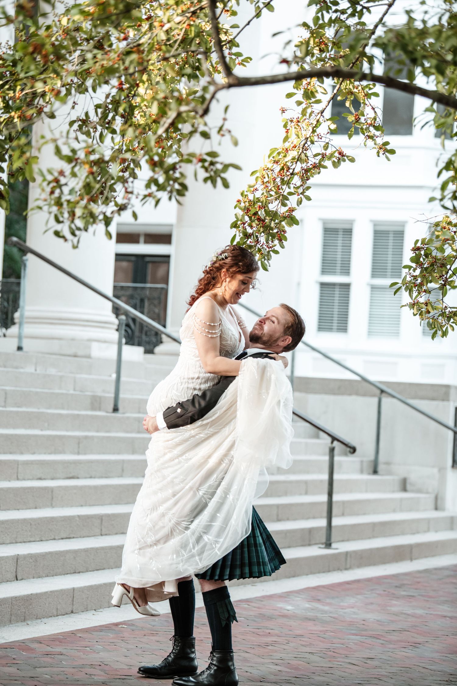 Playful wedding couple dancing and twirling on brick pavement with trees overhead.