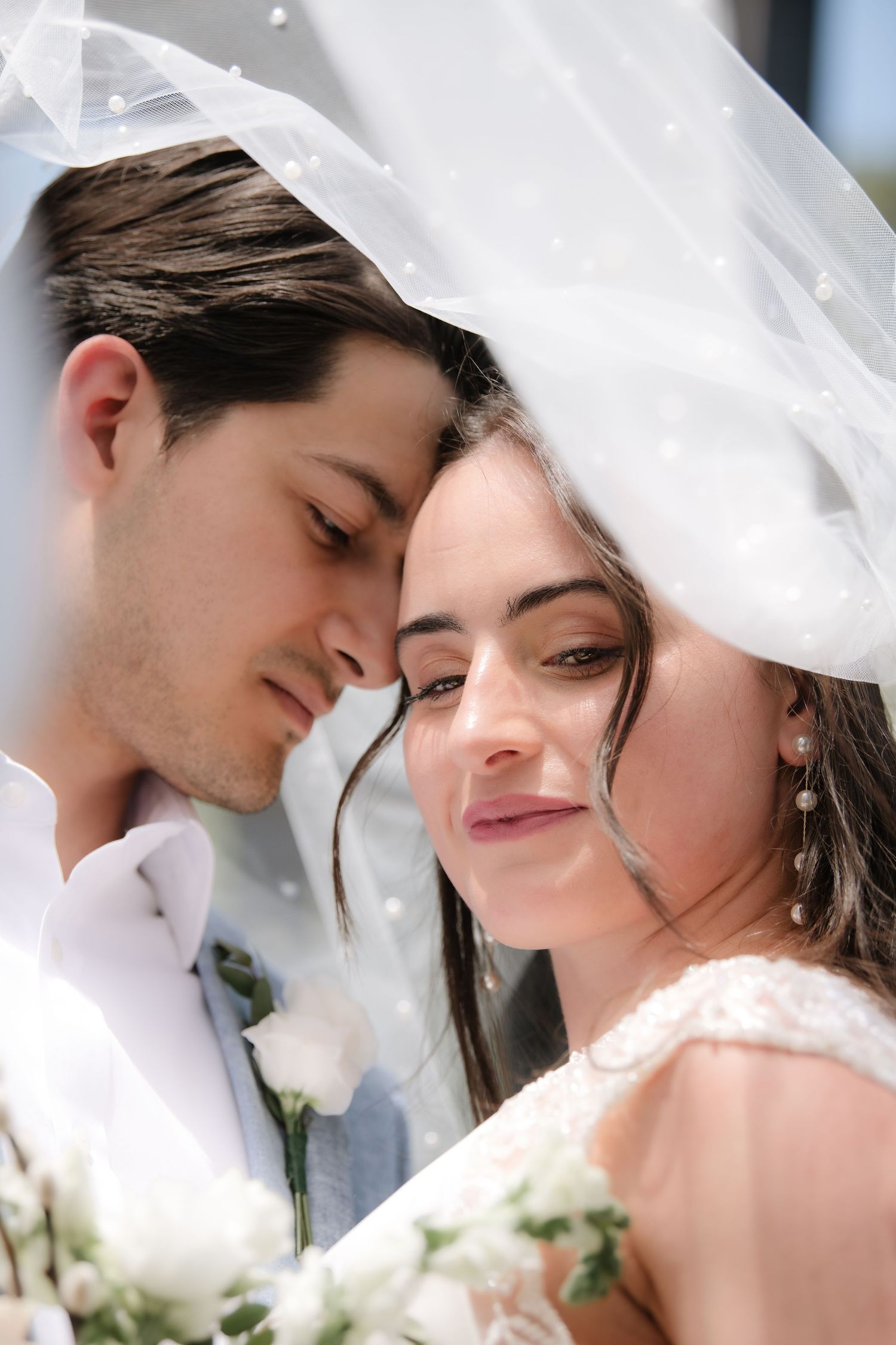 Romantic close-up wedding portraits of a couple under a white veil with soft lighting and white floral accents.