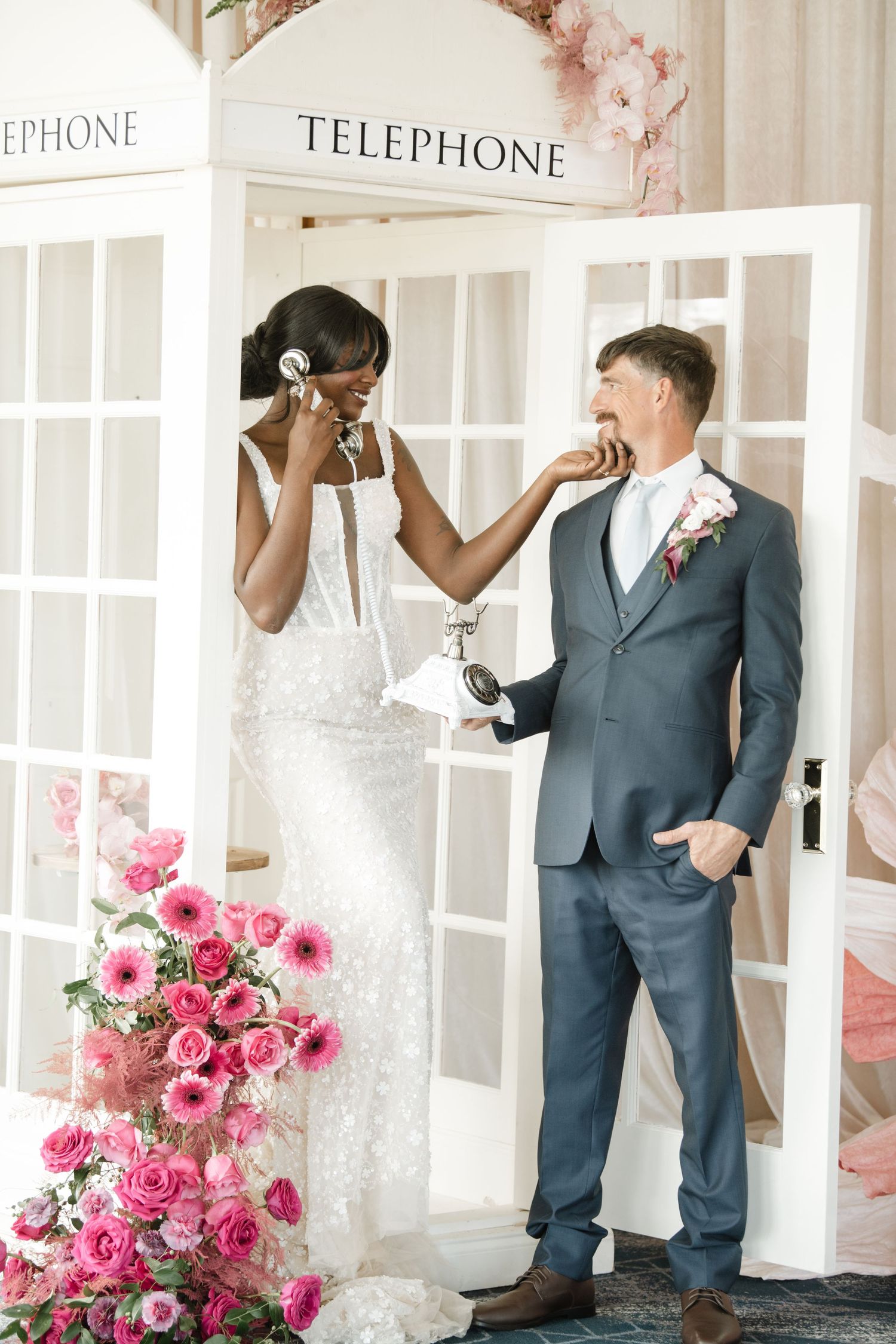 Couple poses playfully with vintage telephone booth and pink floral arrangements.