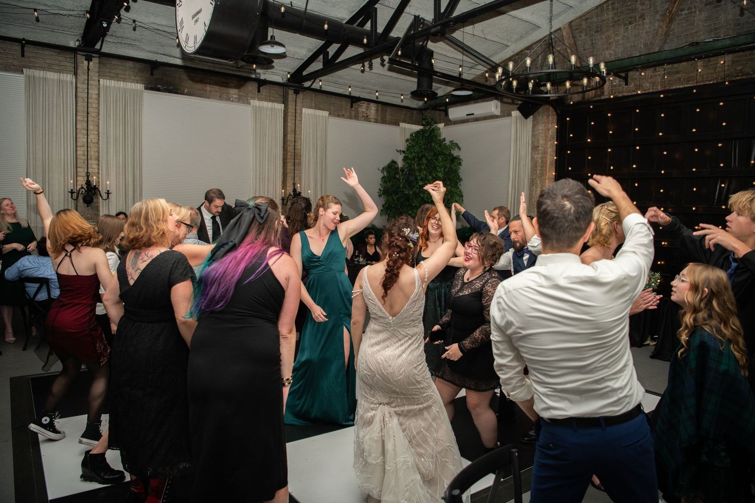 Wedding party dancing with raised arms under string lights at indoor reception.