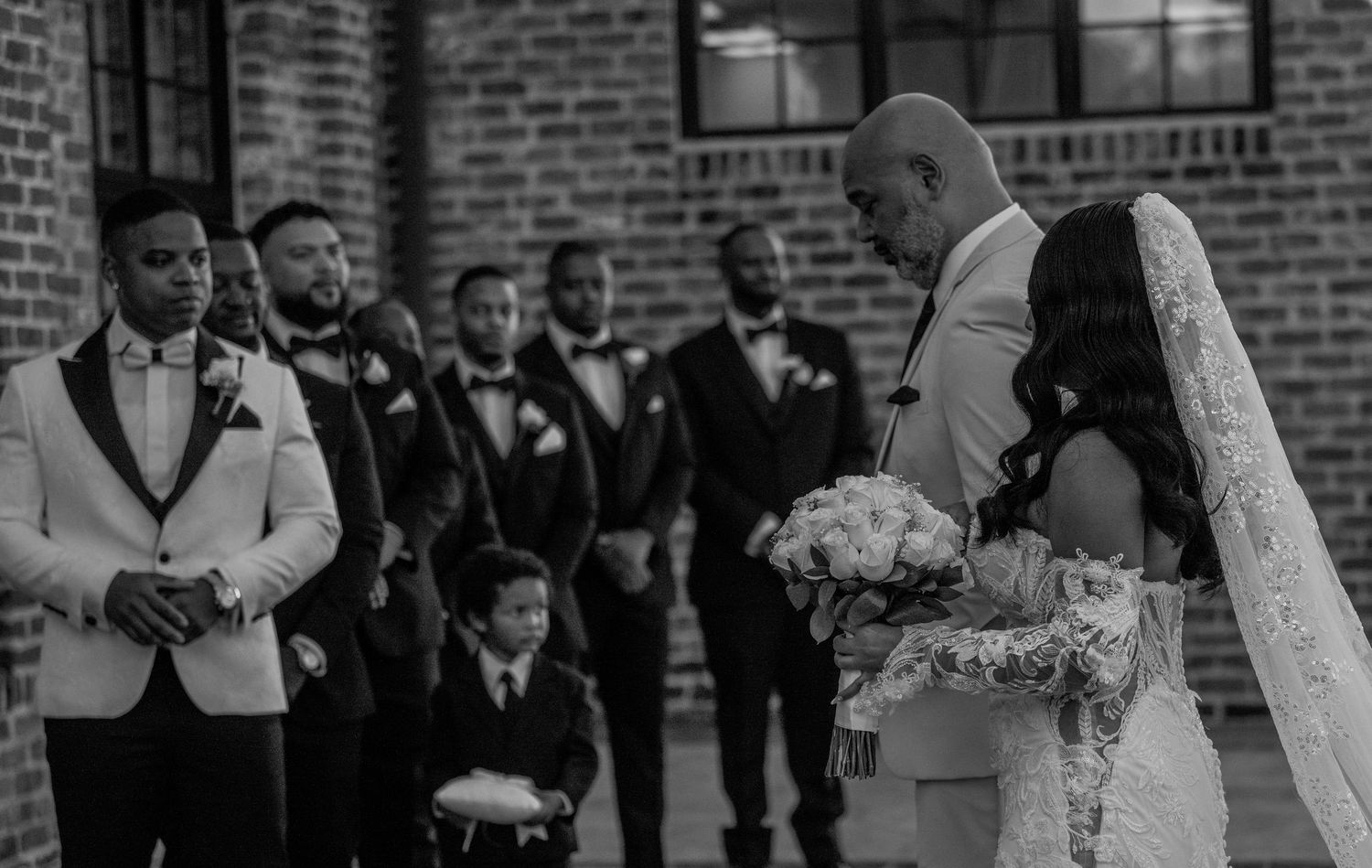 A black and white wedding photo showing the wedding party gathered outside a brick building.