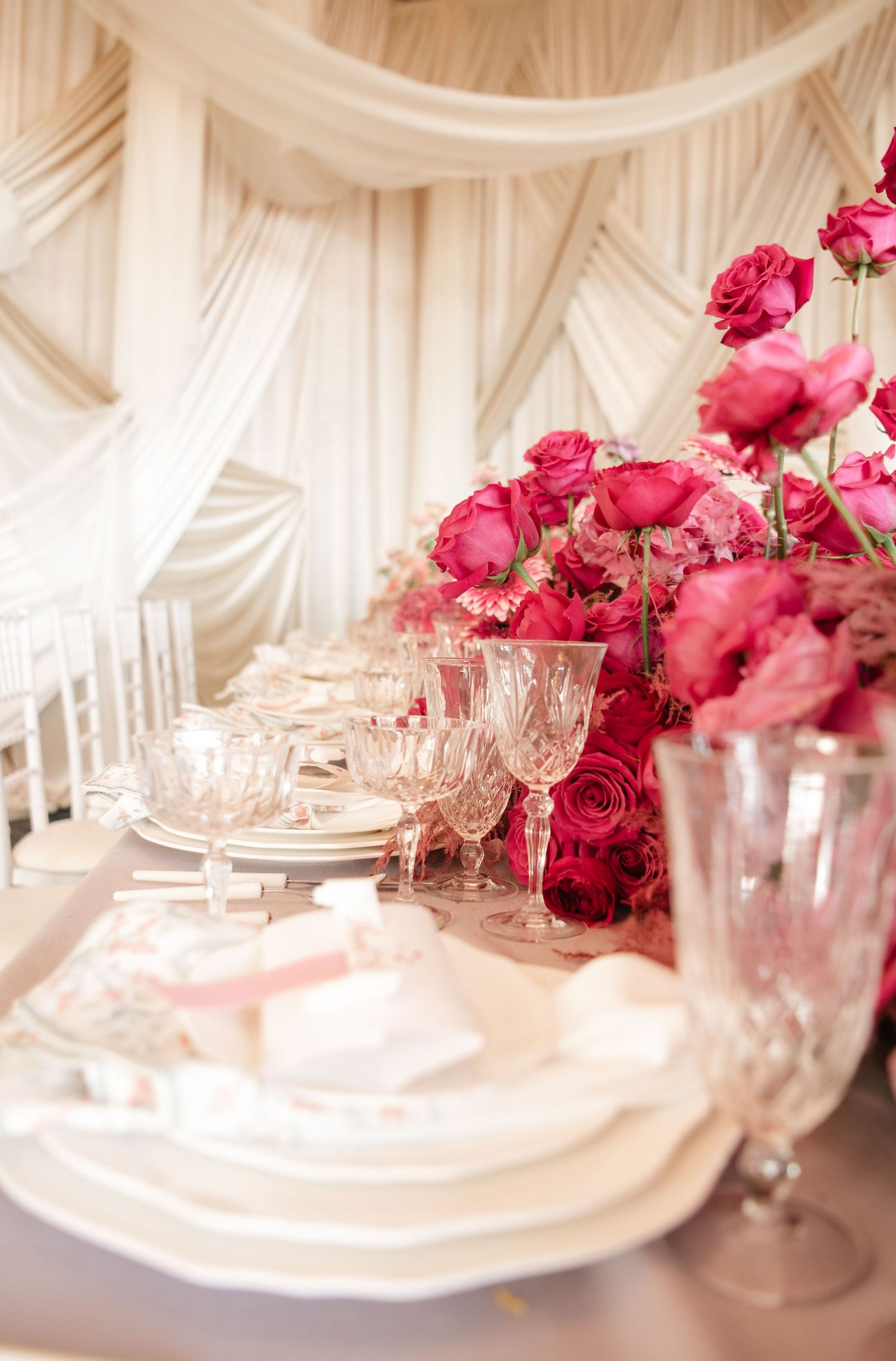 Luxurious wedding table setting with pink roses and crystal glassware against white draping.