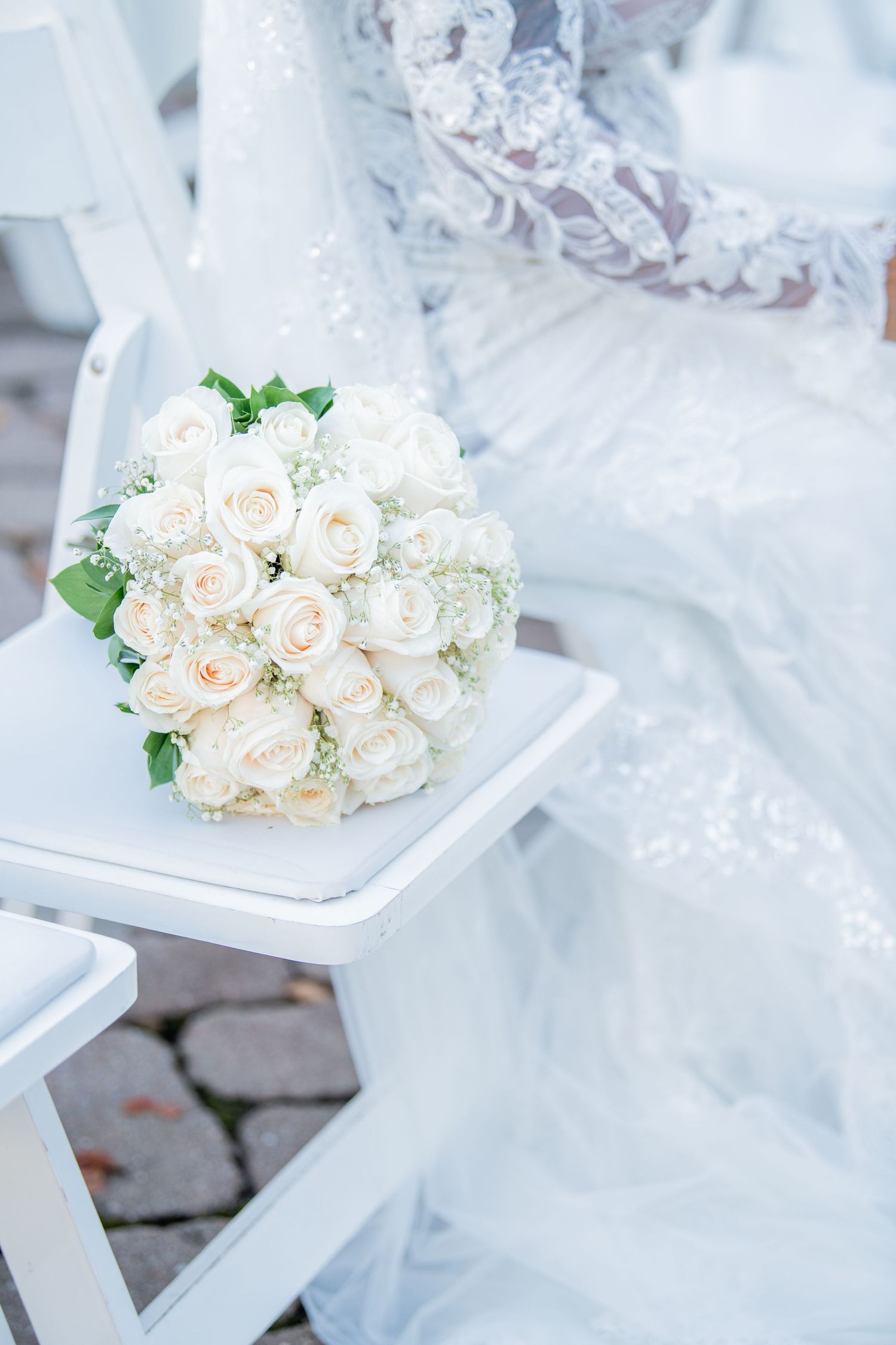 Delicate white rose wedding bouquet resting on white chair against soft background.