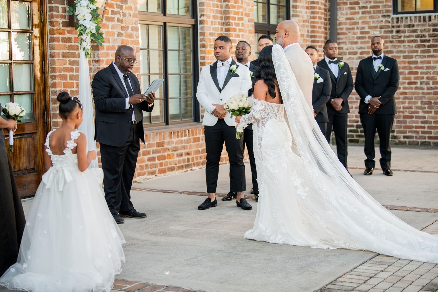 Wedding party gathers for photos in front of rustic brick building with wooden doors.