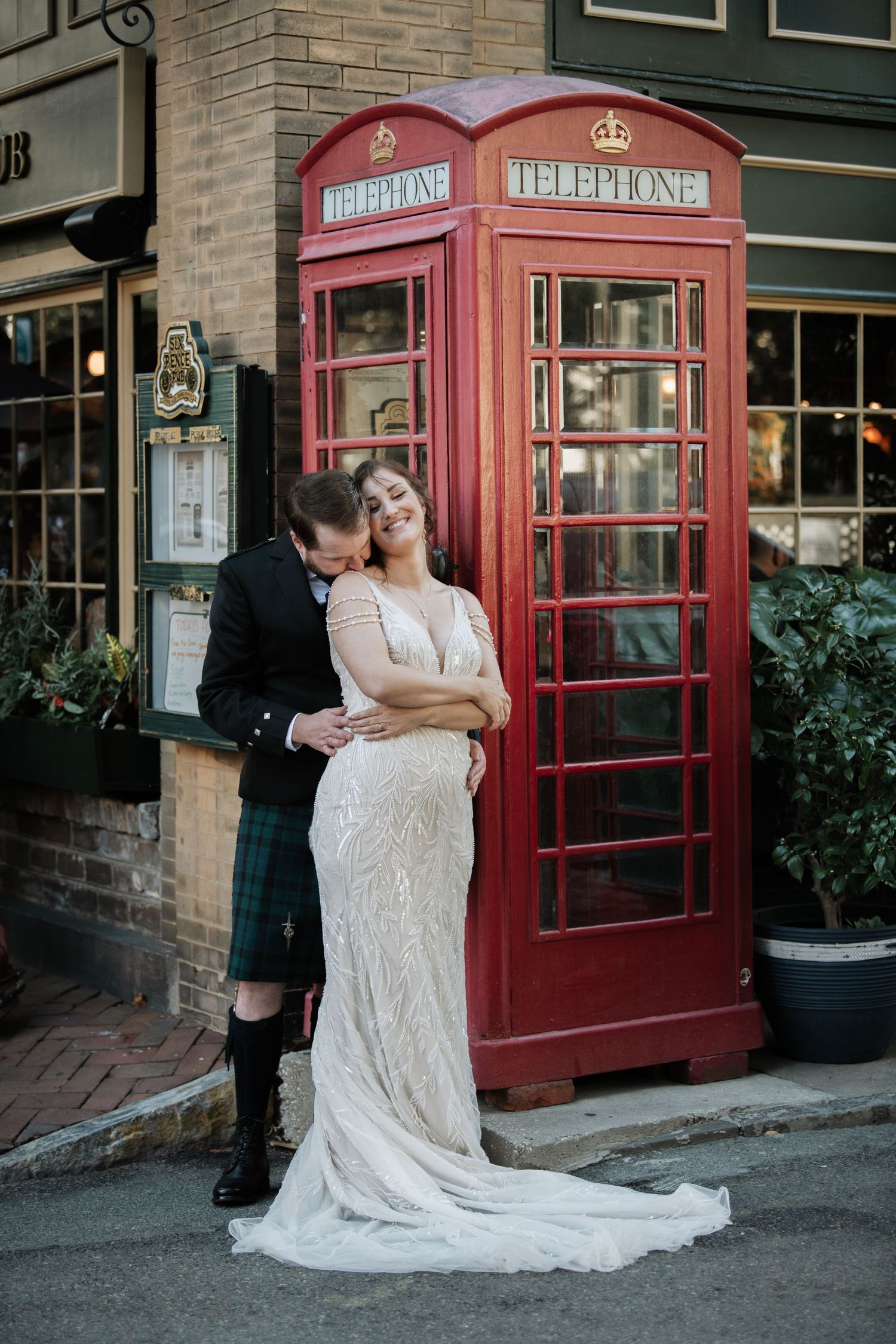 Wedding couple embracing near red British phone boxes on a brick street.