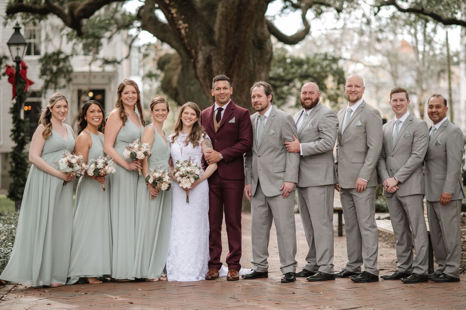 Wedding party in sage green and gray attire poses outdoors under Spanish moss trees.