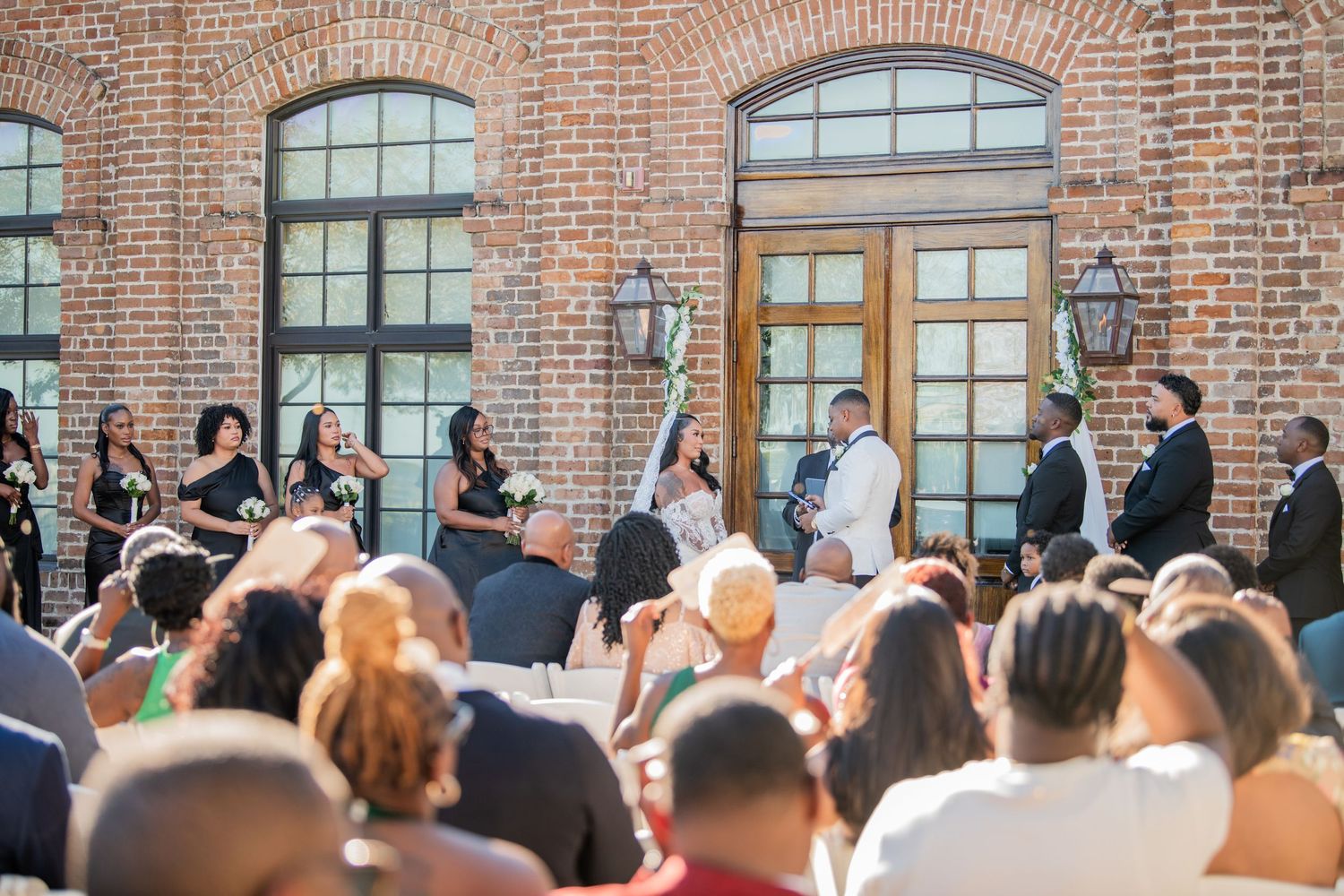 Wedding ceremony attendees line up along a rustic brick wall with arched windows.