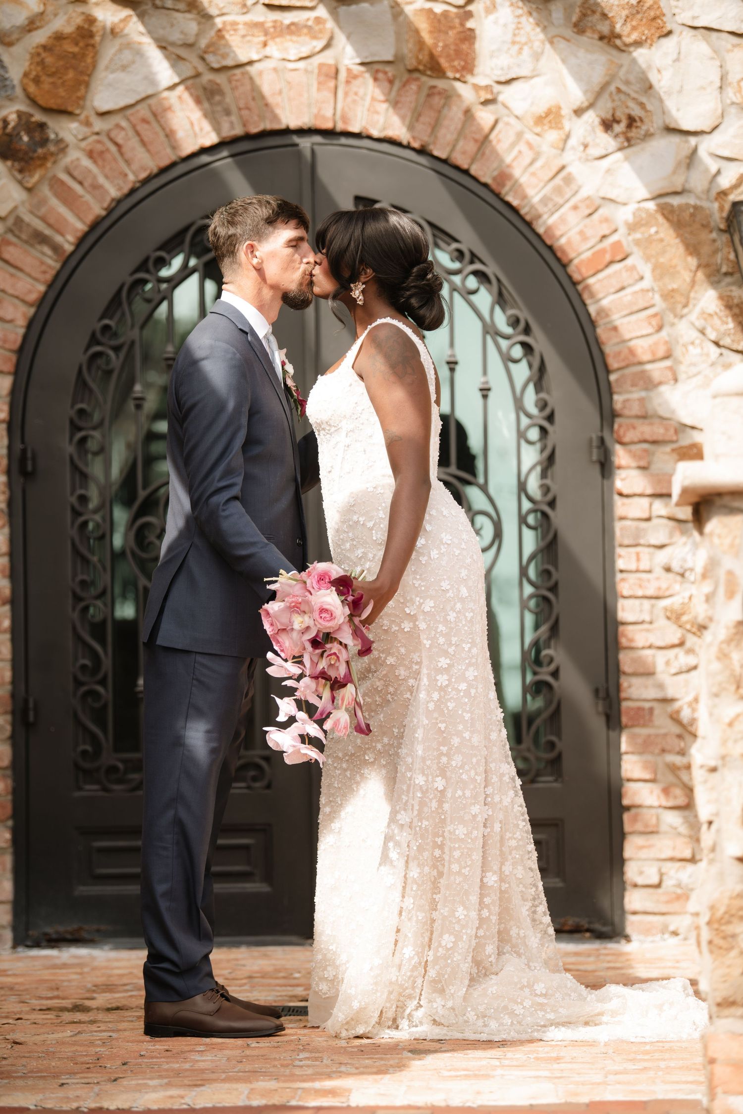 A romantic wedding couple shares an intimate moment under a stone archway with wrought iron details.