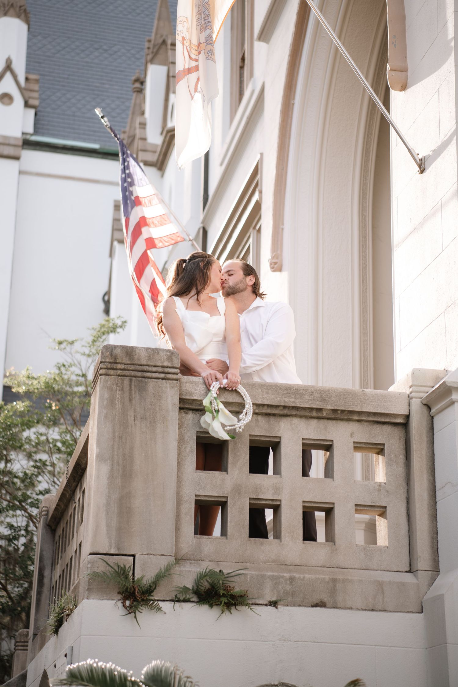 A romantic couple shares a tender moment on a stone balcony with an American flag waving in the background.