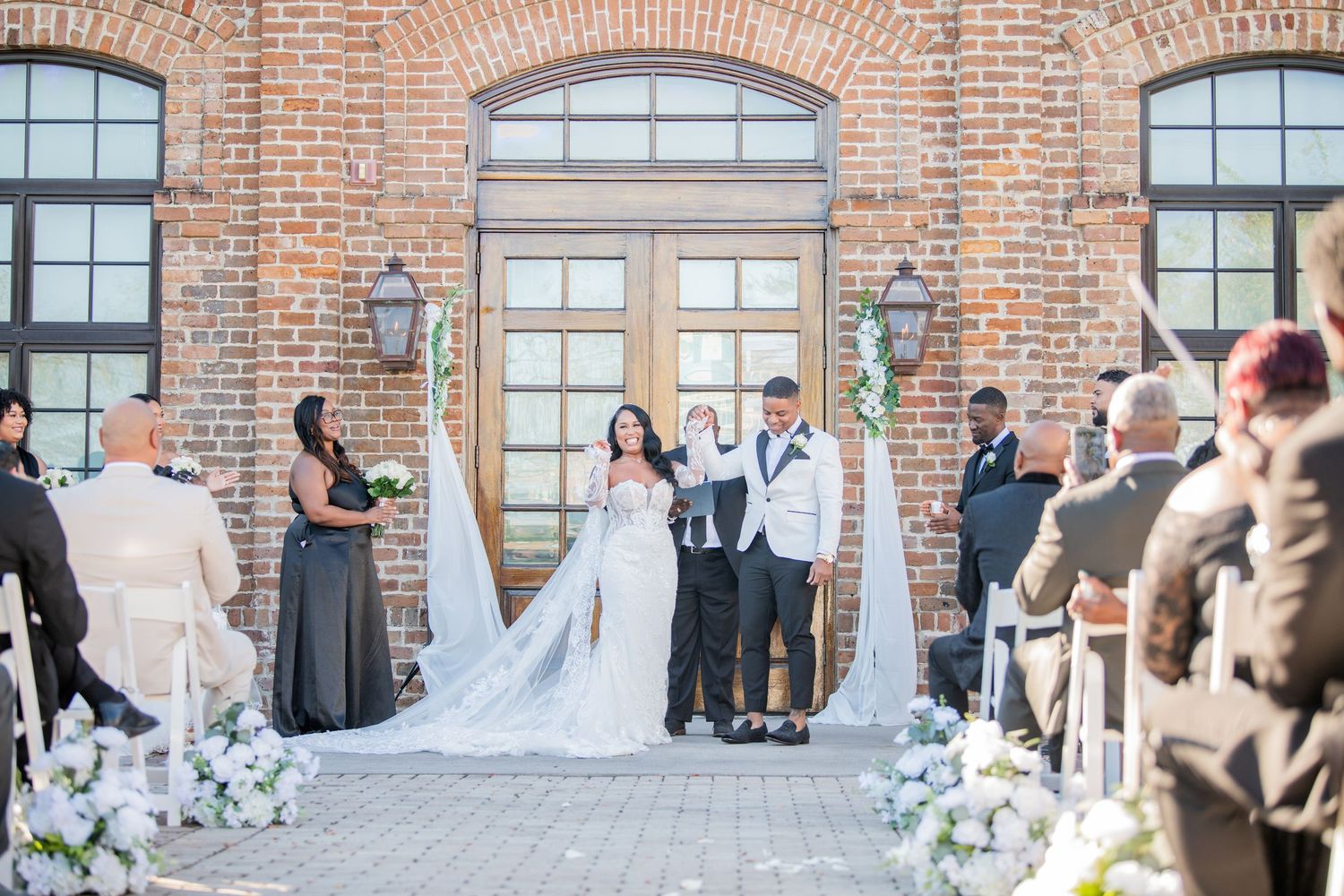 Wedding party and guests celebrating outside historic brick venue with floral decorations.