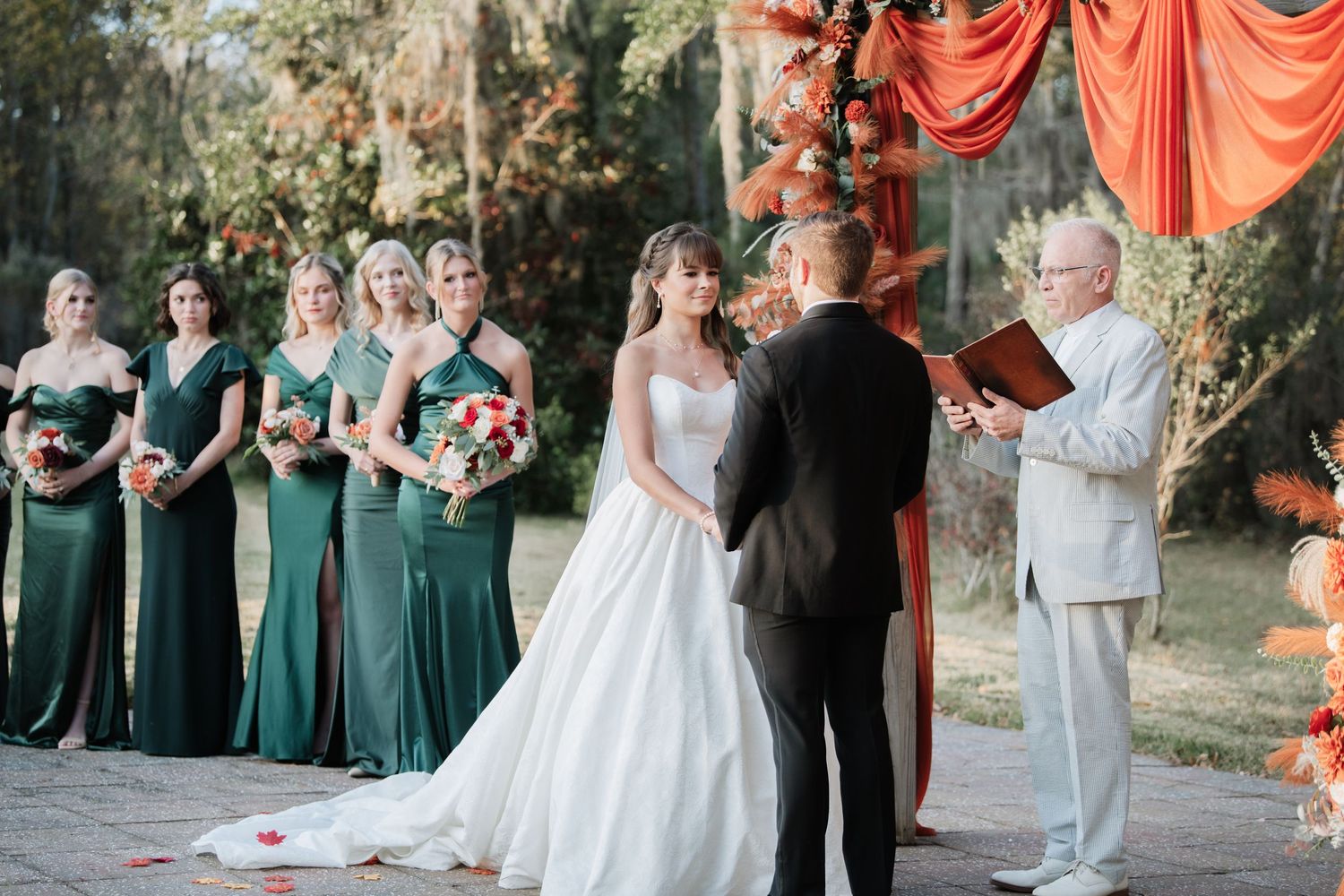Wedding ceremony with bridesmaids in emerald dresses and coral fabric decor.