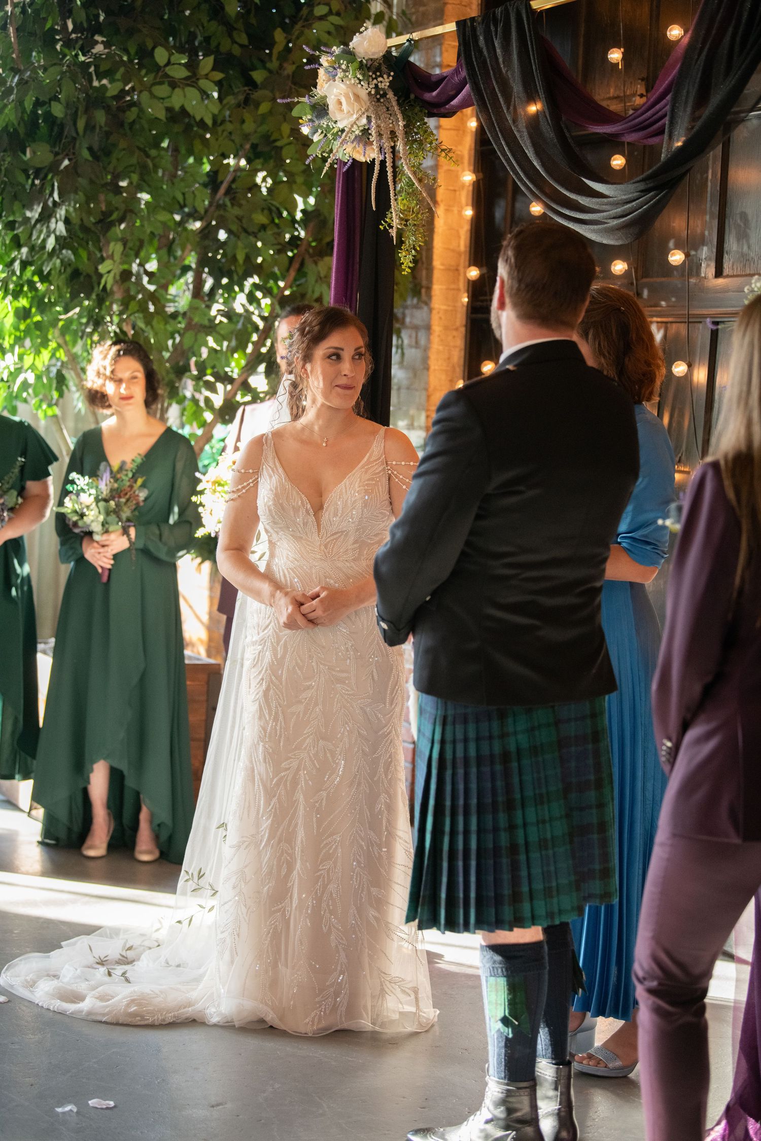 Wedding guests gather in a rustic venue decorated with greenery and string lights.