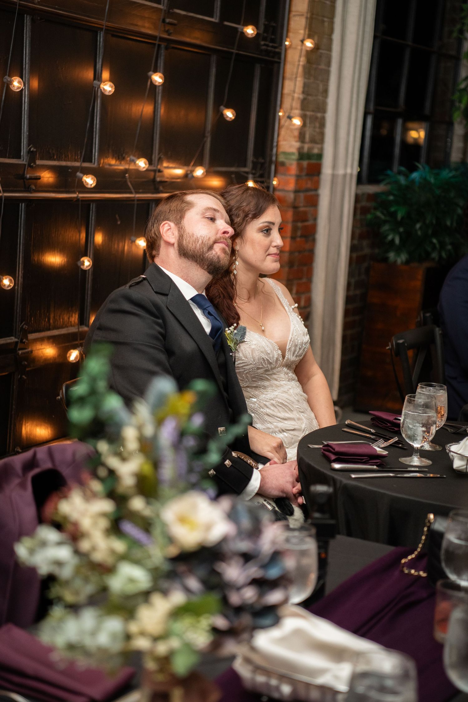 Wedding reception table setting with purple linens and floral centerpieces under string lights.